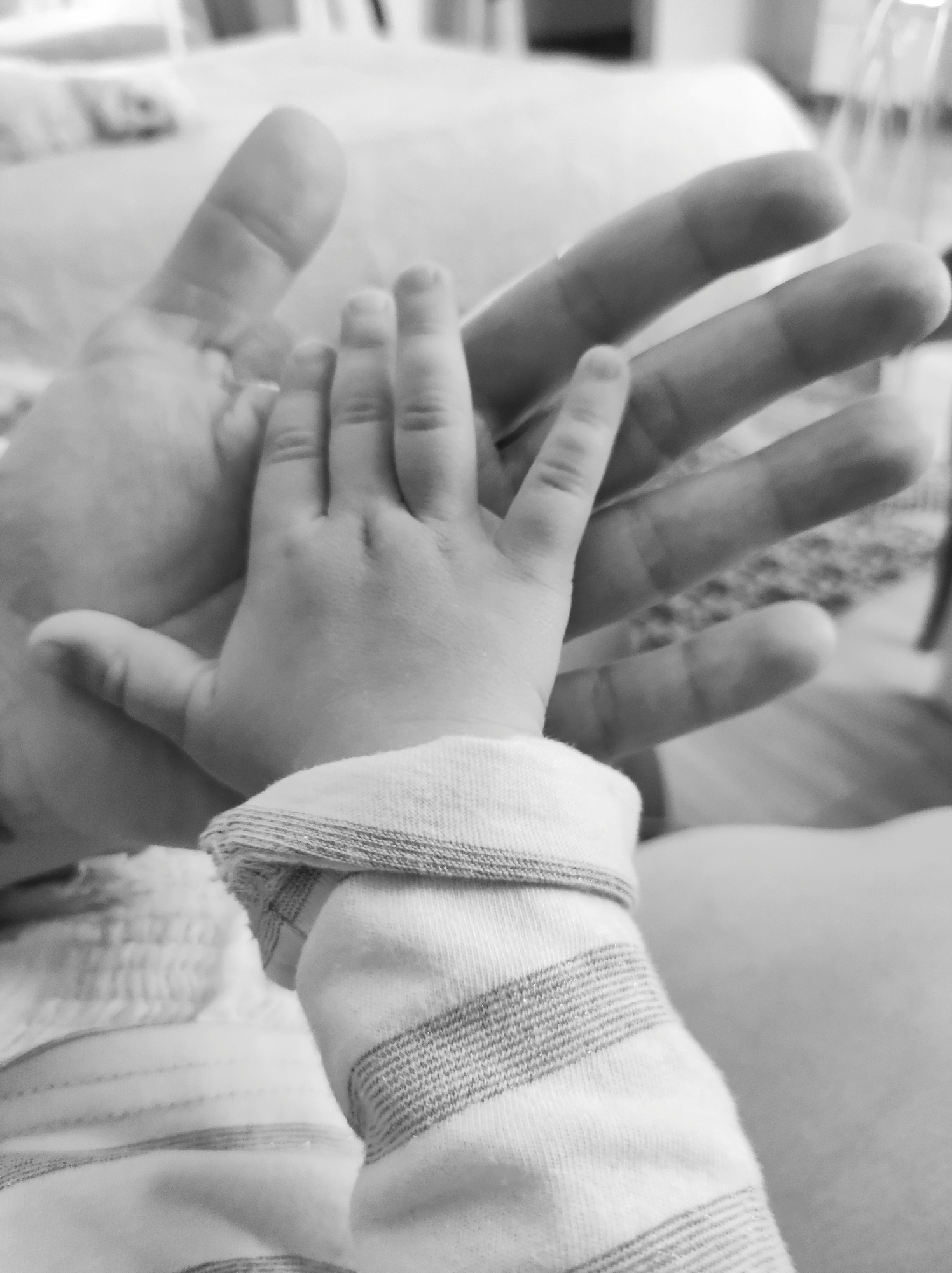 A black and white photograph depicts a child's hand resting on an adult's open hand. The child's hand, clad in a striped, long-sleeved garment, has fingers spread against the adult's palm. The adult's hand is larger, with elongated fingers, and is slightly out of focus. The background is blurred, but some furniture like a bed and a chair are visible. There is no information to suggest the location is in Фоки, Russia.