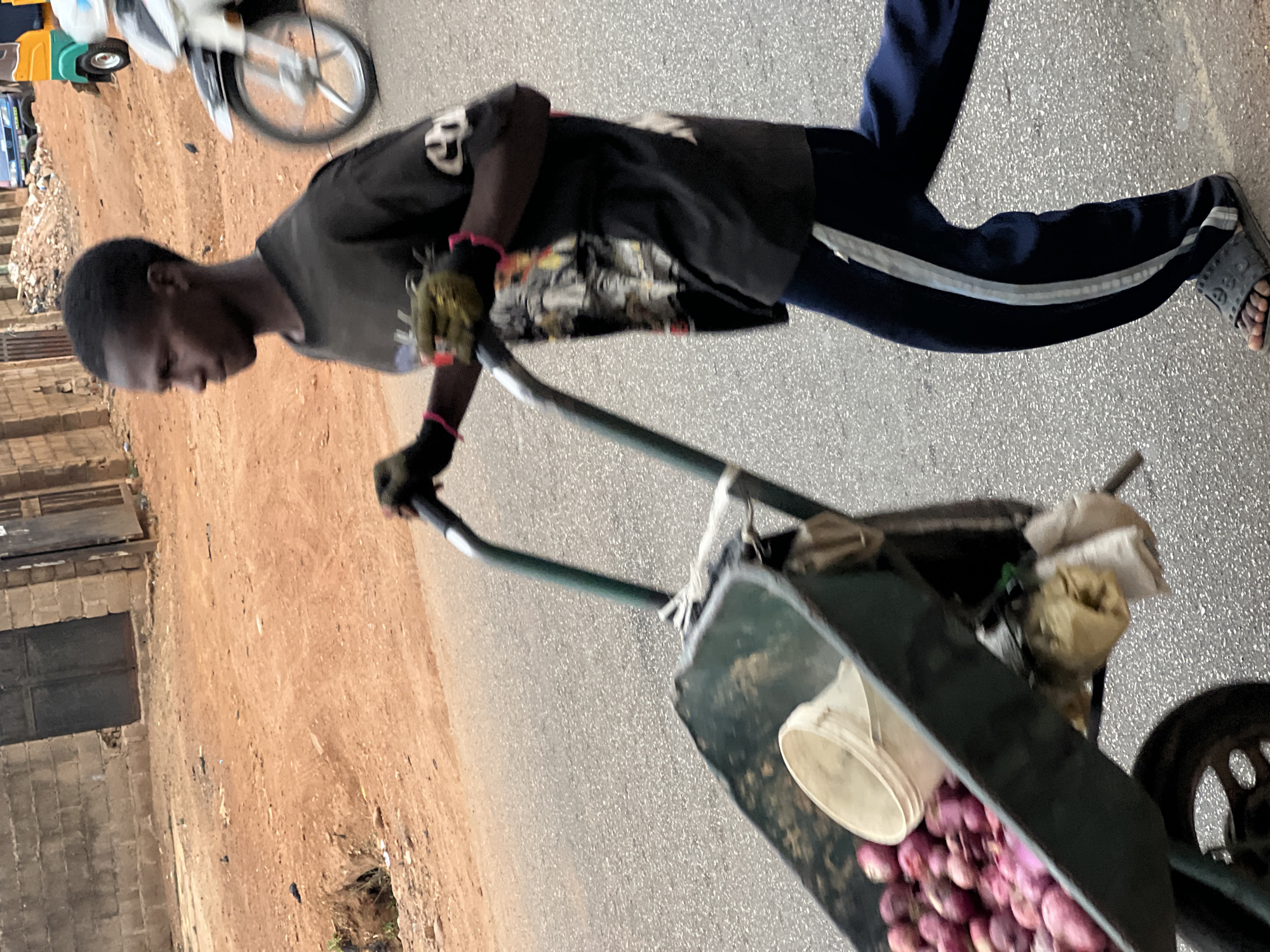 A man is pushing a wheelbarrow full of red onions on a paved road. He is wearing a black t-shirt, navy pants with a white stripe, gloves, and sandals. The wheelbarrow is green and also contains a white bucket and some bags. The background shows a dirt road and some buildings. A motorcycle is parked nearby. The scene is in Gura Top, Nigeria.