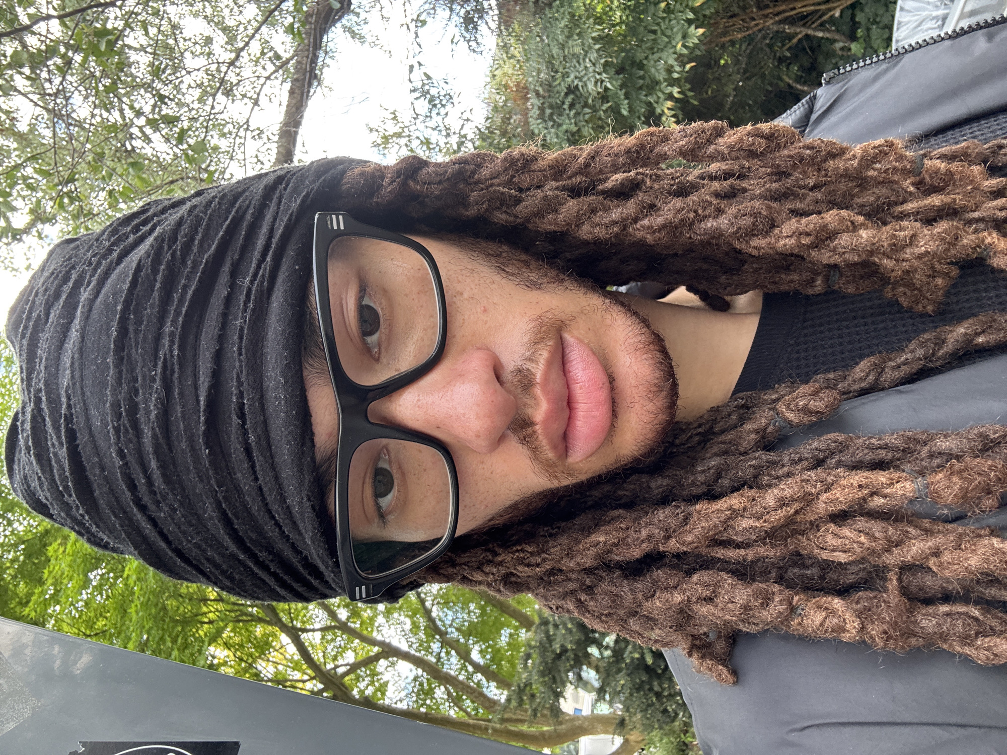 A man wearing a black head covering and black-rimmed glasses is looking directly at the camera. He has brown dreadlocks that frame his face. He is wearing a black shirt and a grey jacket. In the background are trees, a bit of sky, and what appears to be the corner of a building. The location is likely Les Ulis, France, based on the user's request, although there are no distinct landmarks that would confirm the exact location within that city.