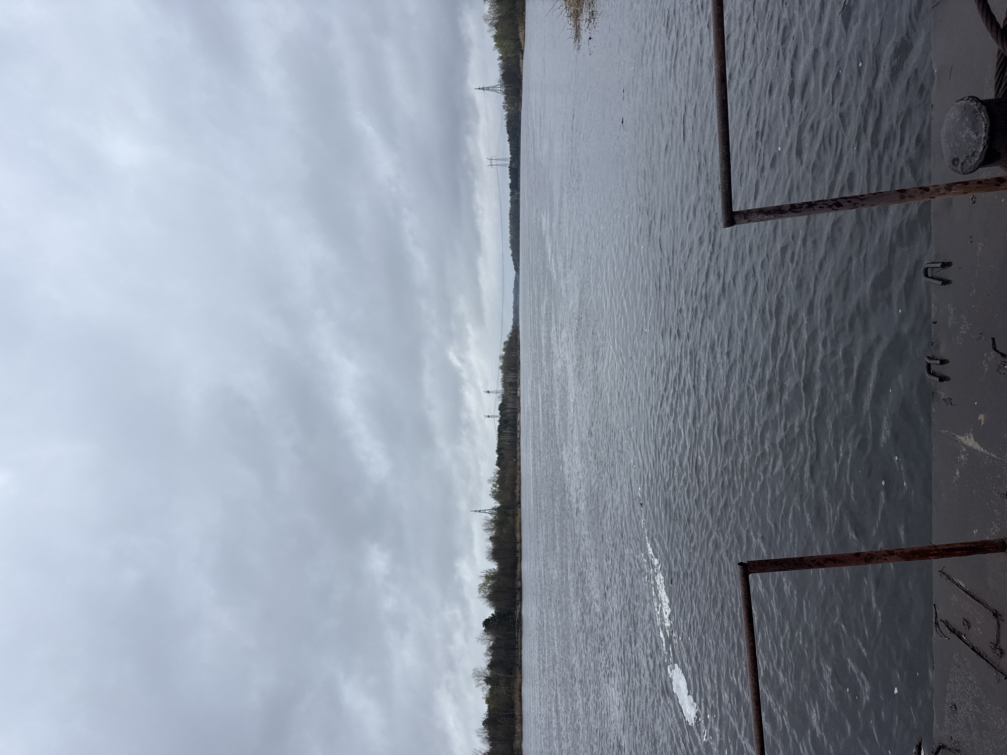 The image shows a body of water, likely a river or lake, with visible ripples and dark, gray coloration. The sky is overcast, with layers of gray clouds. Trees and power lines can be seen along the far bank, indicating a potentially industrial or developed area. The right edge of the frame includes part of a metallic structure with rusty details, possibly from a dock or boat. Given the prompt, this is likely the Pripyat River in Chornobyl, Ukraine. No people are visible in the image.