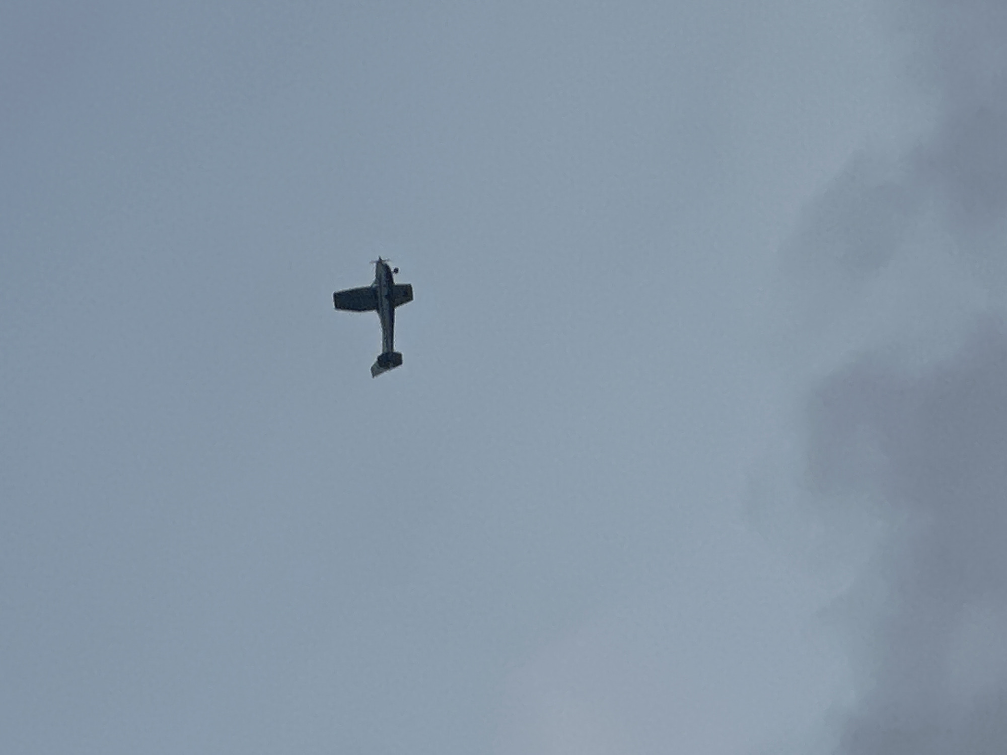 A single, small, propeller-driven airplane is captured in mid-flight against a pale, overcast sky. The aircraft, which appears to be a Cessna or similar light aircraft, is silhouetted against the light sky and is positioned in the upper right quadrant of the image. The plane is oriented with its nose pointing slightly upwards and to the left. The sky is uniformly pale blue-gray, with no discernible landmarks or distinguishing features, suggesting an aerial view high above any specific urban landscape. The lower portion of the image features diffuse, hazy clouds, obscuring any ground-level details. There is no indication of the city of Haifa, Israel, or any specific location within it.