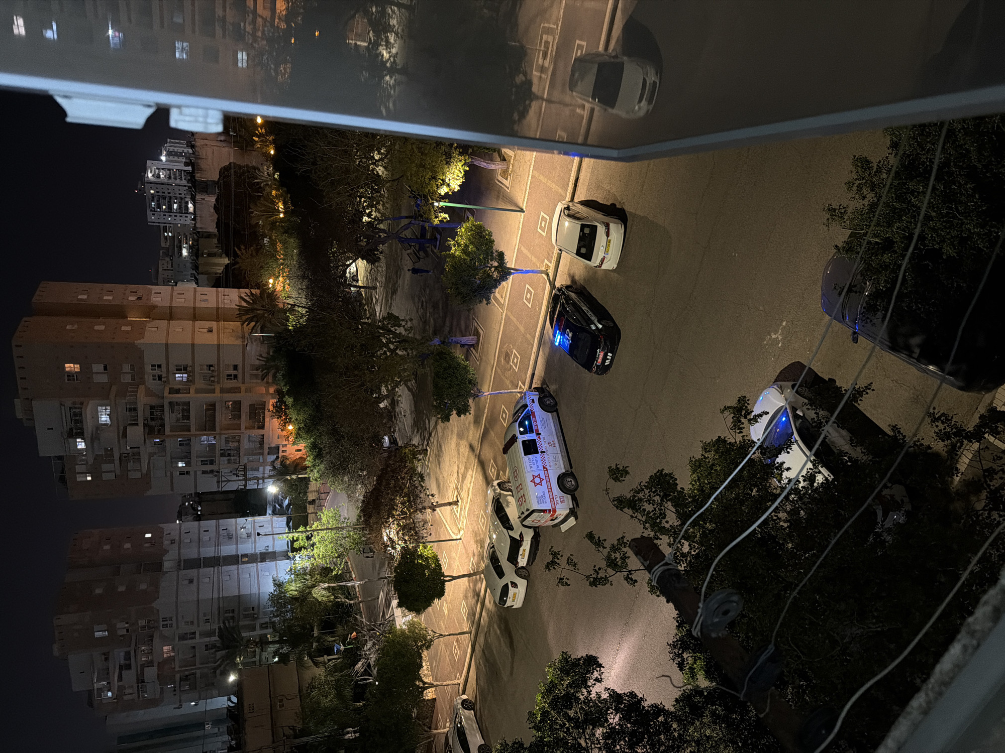 An elevated night-time view captures an urban street scene in Kiryat Bialik, Israel, featuring emergency vehicles. The image is taken from a building, looking down onto a paved road lined with trees and multi-story residential buildings.
The focal point is a white ambulance and a black police car parked on the street, both with emergency lights active. The ambulance, positioned towards the center-right, displays flashing red and blue lights. Its side is marked with the red Star of David emblem and Hebrew text, including "מגן דוד אדום" (Magen David Adom), the emergency number "101", and "1573". The black police car is parked nearby with blue emergency lights on its roof, and partially visible Hebrew text, likely "משטרה" (Police), on its side. Several other white cars are parked along the street or visible in the background.
The street itself features distinctive square-patterned markings for parking spaces. To the left, a playground area with blue structures is dimly lit. Streetlights illuminate the scene, casting shadows from the numerous trees and bushes that line the road. In the background and to the sides, residential apartment buildings show lights in various windows.
The foreground includes a reflection of a white car on a horizontal surface, possibly a balcony railing or window. Utility poles with wires are visible on the right side of the frame. The scene is dark, characteristic of night, with artificial lights providing illumination for the emergency activity. No people are distinctly visible interacting with the vehicles.