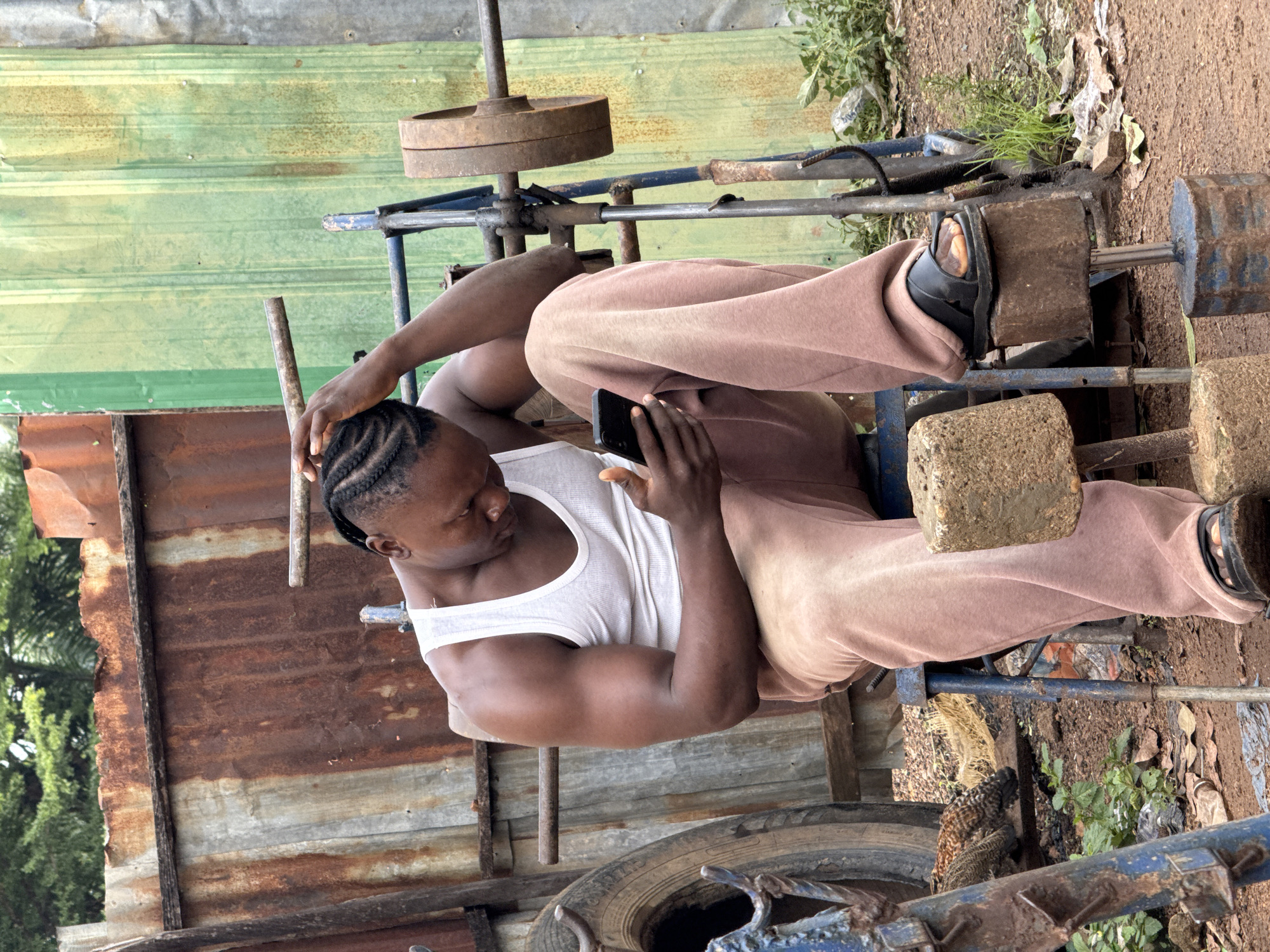 A young Black man sits on a weight bench, holding a smartphone in his right hand, his left arm resting on his head. He wears a white tank top and loose, light brown sweatpants. The bench appears to be a makeshift setup, with rusted metal bars and weights, including a barbell with two large, circular weights. In the background, there are corrugated metal structures, one with a greenish hue and another with significant rust. To the left of the bench is a large tire. The ground is covered in dirt and fallen leaves. This scene is situated in a rugged outdoor environment, likely a gym or training area, within the city of Akwanga, Nigeria.