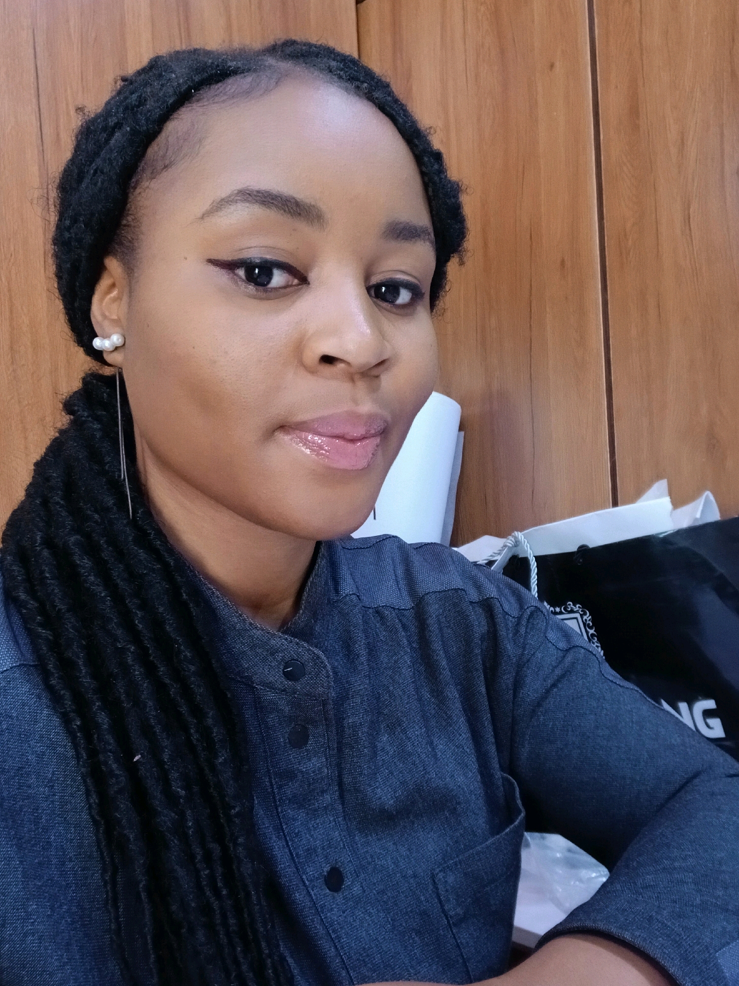 A medium shot captures a young Black woman with braided hair styled in locs, wearing a dark denim collared shirt. She is seated in what appears to be an indoor office environment. The background features wooden paneling on the right and a white surface on the left, possibly a desk or shelf. A black shopping bag with white lettering is visible behind her. The lighting suggests an indoor setting. There is no indication of the specific location within Gura Top, Nigeria.