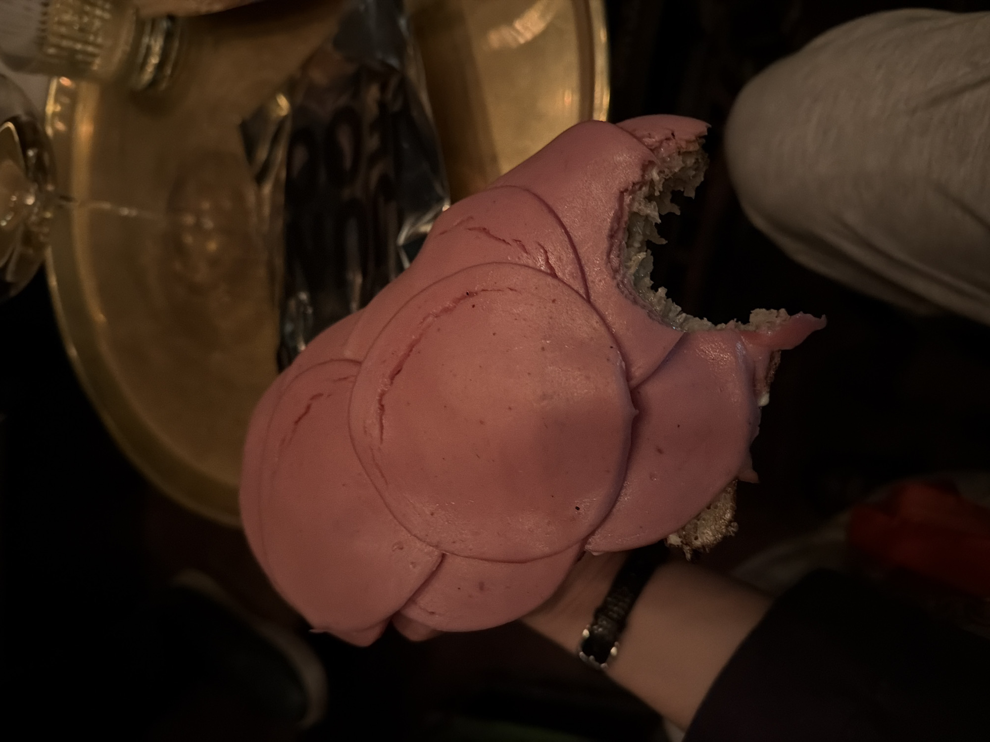 A close-up shot shows a partially eaten sandwich, heavily layered with bologna-like cold cuts, held in someone's hand. The sandwich has a bite taken out of it. A watch is visible on the wrist of the person holding the sandwich. In the background, a silver tray and a golden-colored drinking vessel are visible. A portion of another person's leg is also visible in the upper right. The image seems to have been taken indoors under low lighting conditions. There is no information available to pinpoint this location in Moscow, Russia.