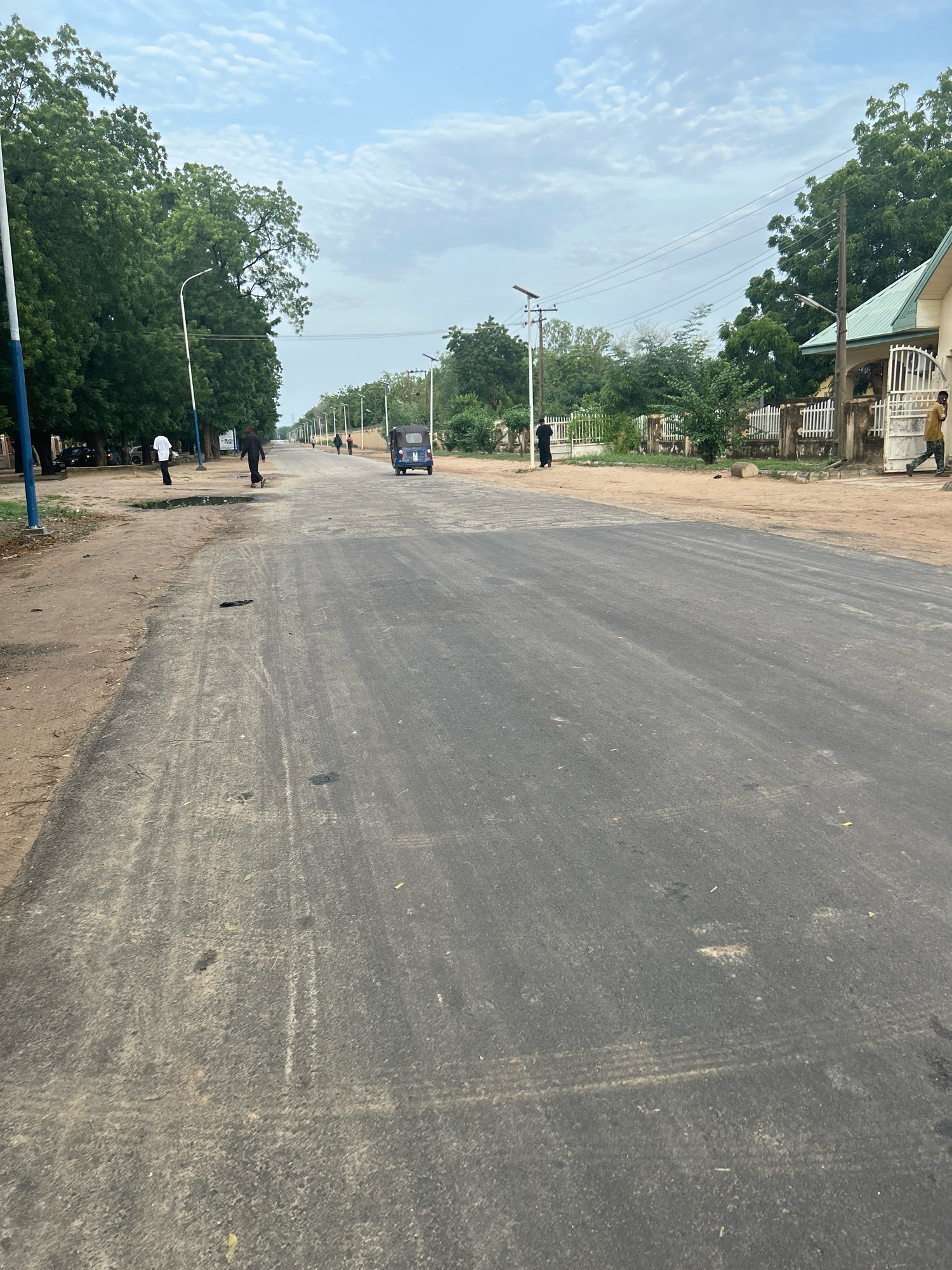 The image shows a paved road in Mairi, Nigeria. A blue auto-rickshaw is parked on the right side of the road. Several people are walking on the road and sidewalk. Trees line both sides of the road, and there are houses and fences visible in the background. The sky is overcast with some clouds. The road appears to be relatively new, with some tire marks visible on the surface.