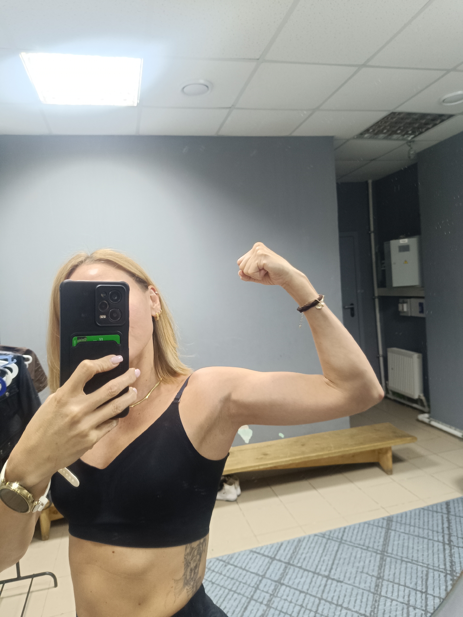 A woman is taking a selfie in a room, likely a gym or locker room. She is wearing a black sports bra and flexing her right bicep. She holds a black smartphone with a green card holder attached to the back. She has blonde hair, a gold necklace, and a gold wristwatch. A light brown bracelet is on her right wrist. Part of a tattoo is visible on her torso. The background shows a light gray wall, a wooden bench, a radiator, and a metal structure with electrical boxes. Based on visual cues, the location appears to be Naberezhnye Chelny, Russia.