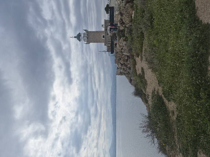 An image depicts a coastal scene at Cape Heraion in the Municipal Unit of Loutraki - Perachora, Greece. A stone lighthouse with a white lantern room and a dark green dome stands prominently on a rocky promontory on the right side of the frame, positioned above a body of water. Approximately four to five individuals are observed standing near the base of the lighthouse, partially obscured by the structure and rocks. The immediate foreground features uneven, sandy-brown terrain interspersed with patches of green vegetation, including small shrubs and what appear to be wildflowers. A calm, gray-blue body of water occupies the left and central midground, extending to a distant, hazy landmass of hills or mountains under an overcast sky filled with numerous gray and white clouds. The overall lighting suggests a muted daylight condition.