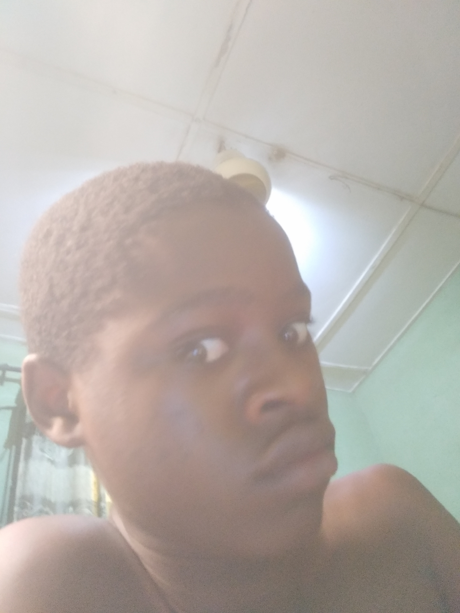 A close-up shot of a person with short hair, light brown skin, a slightly furrowed brow, and a neutral expression. A ceiling fan is visible in the background, mounted on a white ceiling. The location is unclear.