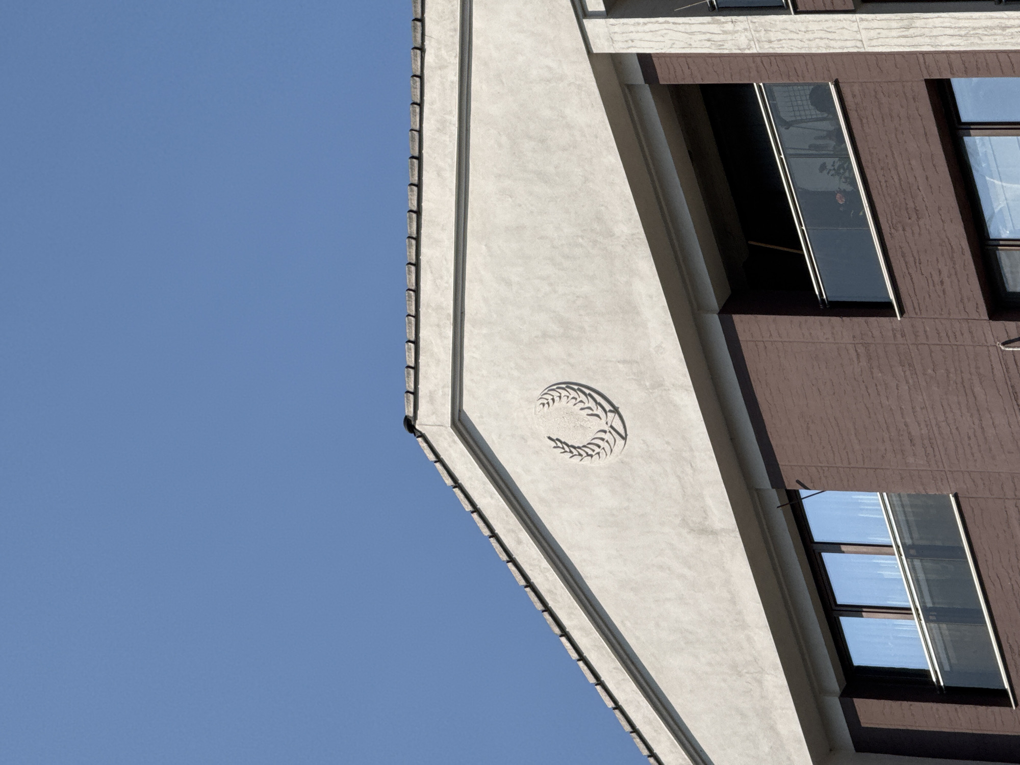 A building with a peaked roof and a stucco facade is visible against a clear blue sky. A circular laurel wreath emblem is embossed on the upper part of the facade. Below the emblem, the building has multiple stories with dark brown brick-like siding. There are large windows with reflective glass on the lower levels and a balcony with a glass railing on the upper level. The scene is likely in the city of Plovdiv, Bulgaria, given the architectural style often seen in modern residential buildings there. No people are visible in the image.