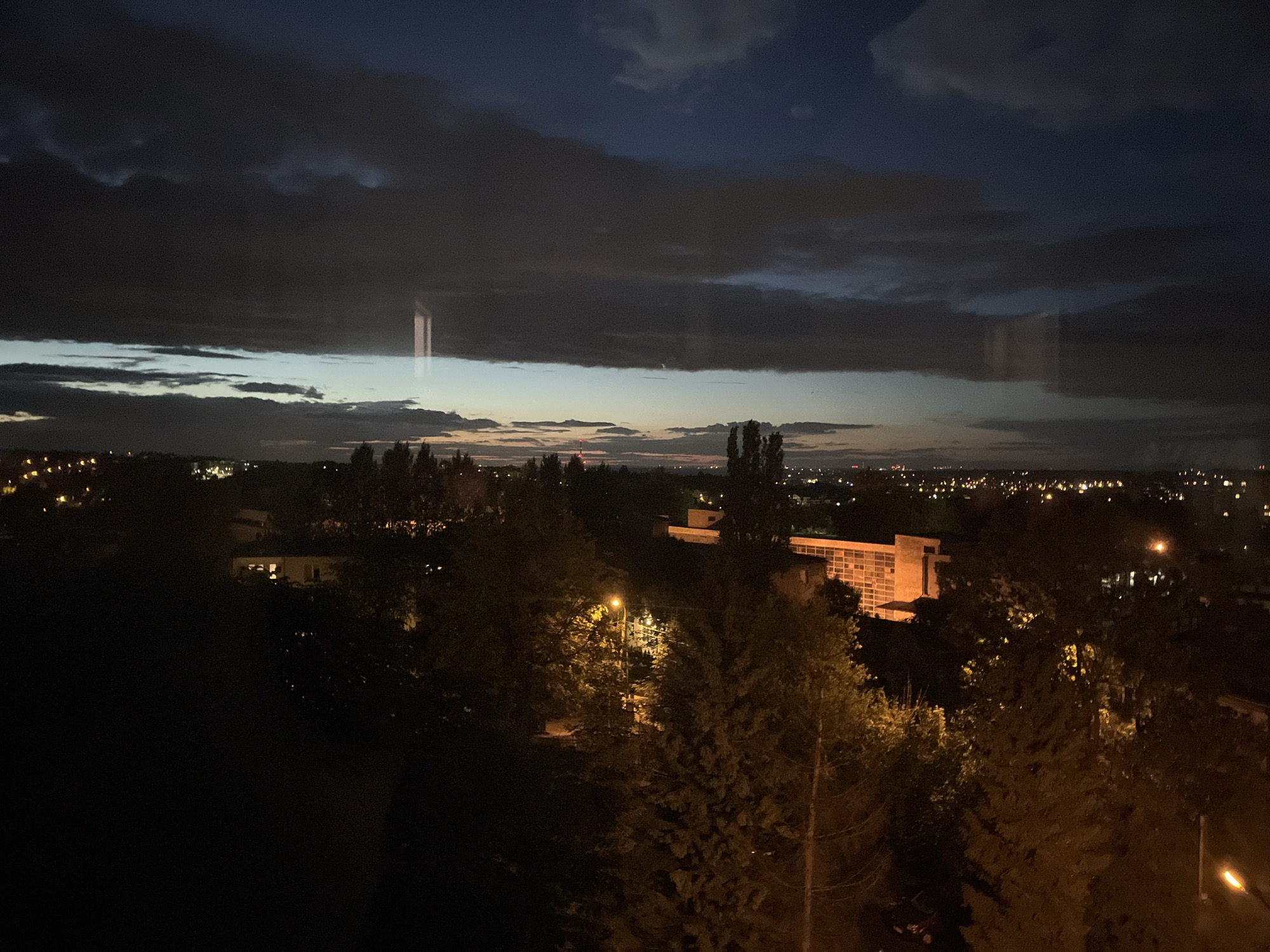 The image is an elevated nighttime view of a city under a dark, cloudy sky. The sky transitions from deep blue at the top to lighter shades of blue and gray near the horizon, with streaks of darker clouds obscuring the upper atmosphere. The city lights twinkle in the distance, suggesting a sprawling urban landscape. In the foreground, silhouetted trees and foliage are prominent, with some illuminated by streetlights. A notable building with illuminated windows is visible in the mid-ground, its facade partially obscured by the trees. Another larger building, possibly an institution or office block with a lighter-colored facade, stands out to the right, also featuring illuminated windows. A streetlamp with a bright yellow glow is also visible, casting light on the surrounding vegetation. The overall impression is a serene, yet somewhat somber, depiction of a city at dusk or after dark.
