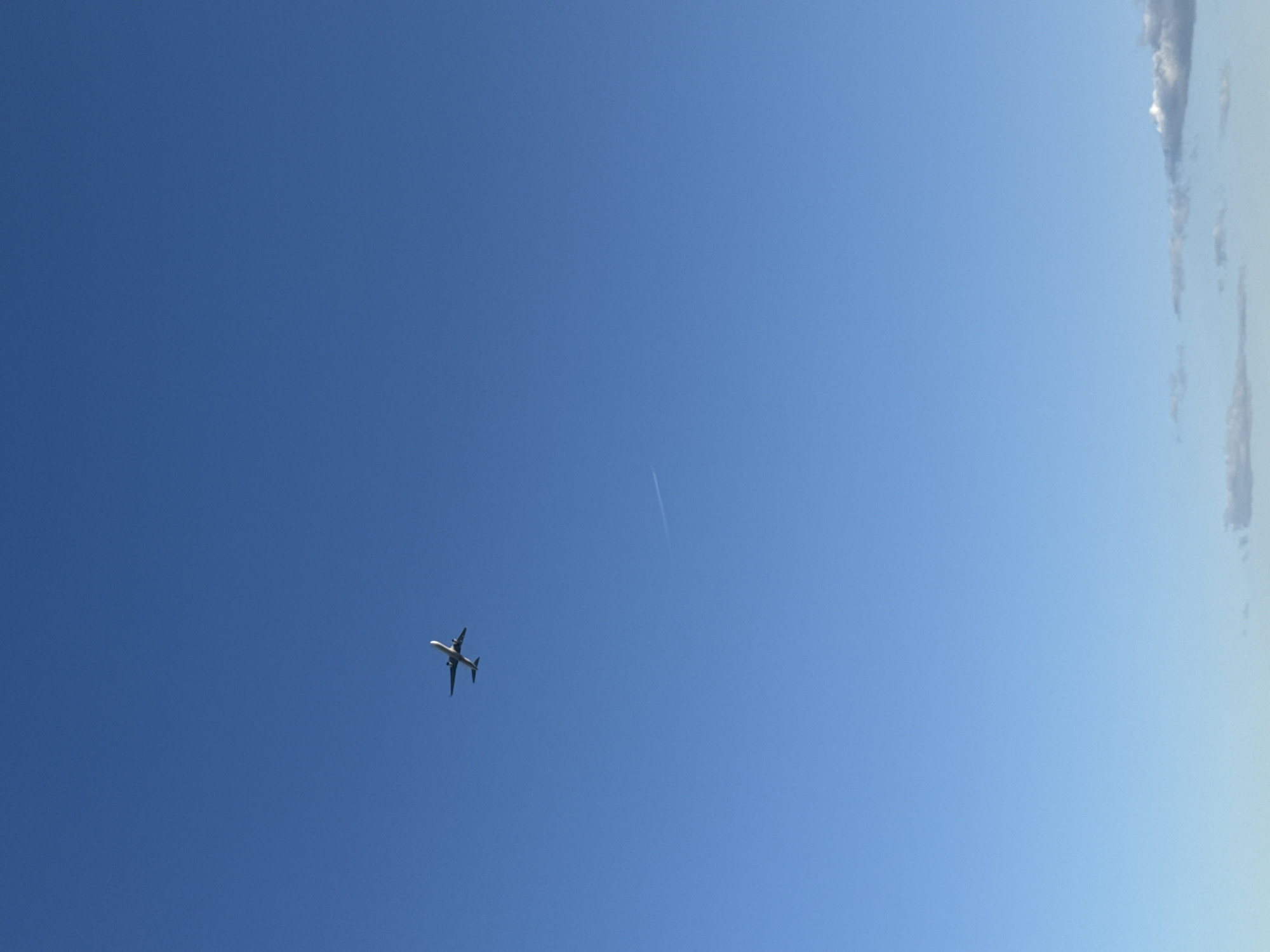 The image shows a clear blue sky with an airplane in the lower left corner. A thin contrail is visible in the upper center of the frame. There are some clouds on the upper right edge of the image. The image does not contain sufficient information to determine a location in Tbilisi, Georgia.