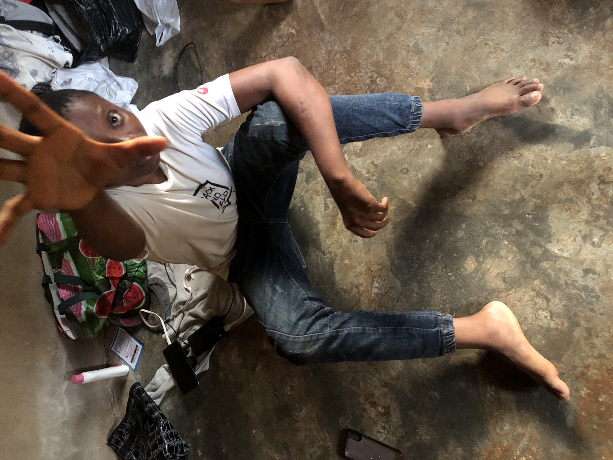 A person lies on a textured, possibly concrete floor of unknown location in an unidentified city. The individual is wearing a white t-shirt with a logo on the chest and blue jeans with the cuffs rolled up. One leg is crossed over the other near the knees. The person's right hand is extended toward the camera. The floor is cluttered with items, including a bag with a watermelon design, a mobile phone, a white tube-shaped object, cables, and an unidentified card. The scene appears to be indoors.