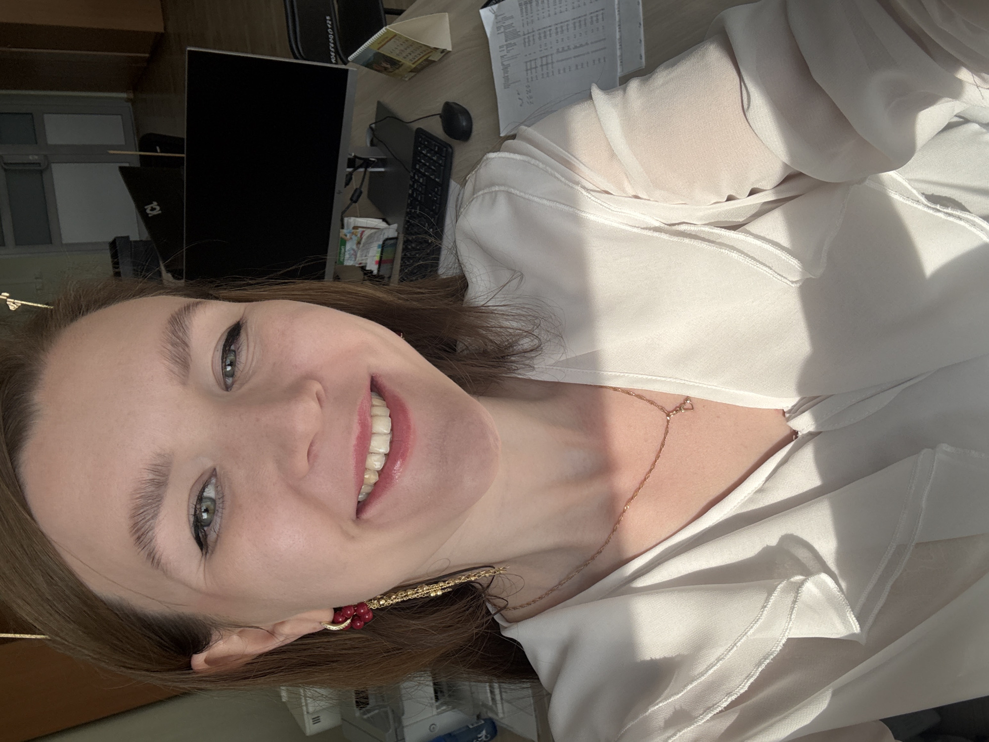 A smiling woman is photographed in a selfie at an office in Izhevsk, Russia. She has fair skin, light brown hair, and is wearing a white blouse with a gold necklace and red earrings. In the background, a computer monitor and keyboard are visible on a desk. There are also papers and office supplies on the desk. Sunlight is shining on the scene.