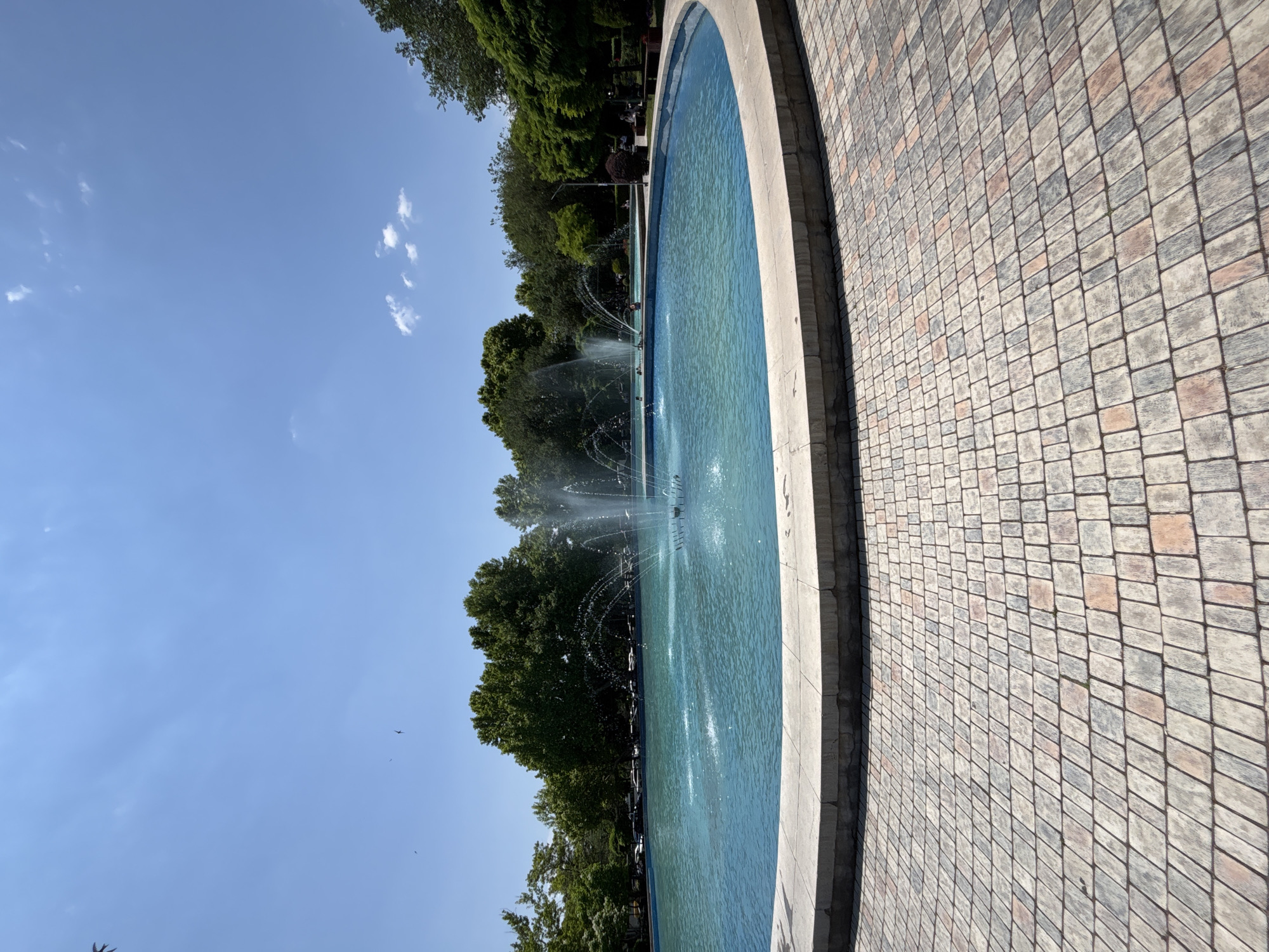 The media file displays a bright, sunny day at a park with a large, circular fountain. The fountain's water is a vibrant turquoise blue, with multiple jets spraying water into the air, creating a lively and refreshing atmosphere. The fountain is surrounded by a light-colored concrete border, and beyond that, a patterned pathway made of square tiles in shades of gray, beige, and reddish-brown.
Lush green trees form a dense backdrop behind the fountain, suggesting a park setting. Some of these trees appear to be mature, with full canopies. In the distance, through the trees, hints of park benches and possibly people can be discerned, though they are not the focus. The sky is a clear, bright blue with a few wispy white clouds, indicating fair weather. The orientation of the image suggests it was taken with the camera held vertically, capturing a wide view of the scene. The overall impression is one of a pleasant, leisurely day in a well-maintained urban park. The time of day appears to be mid-morning or afternoon, given the bright sunlight casting clear shadows. There is no visible text in the image.