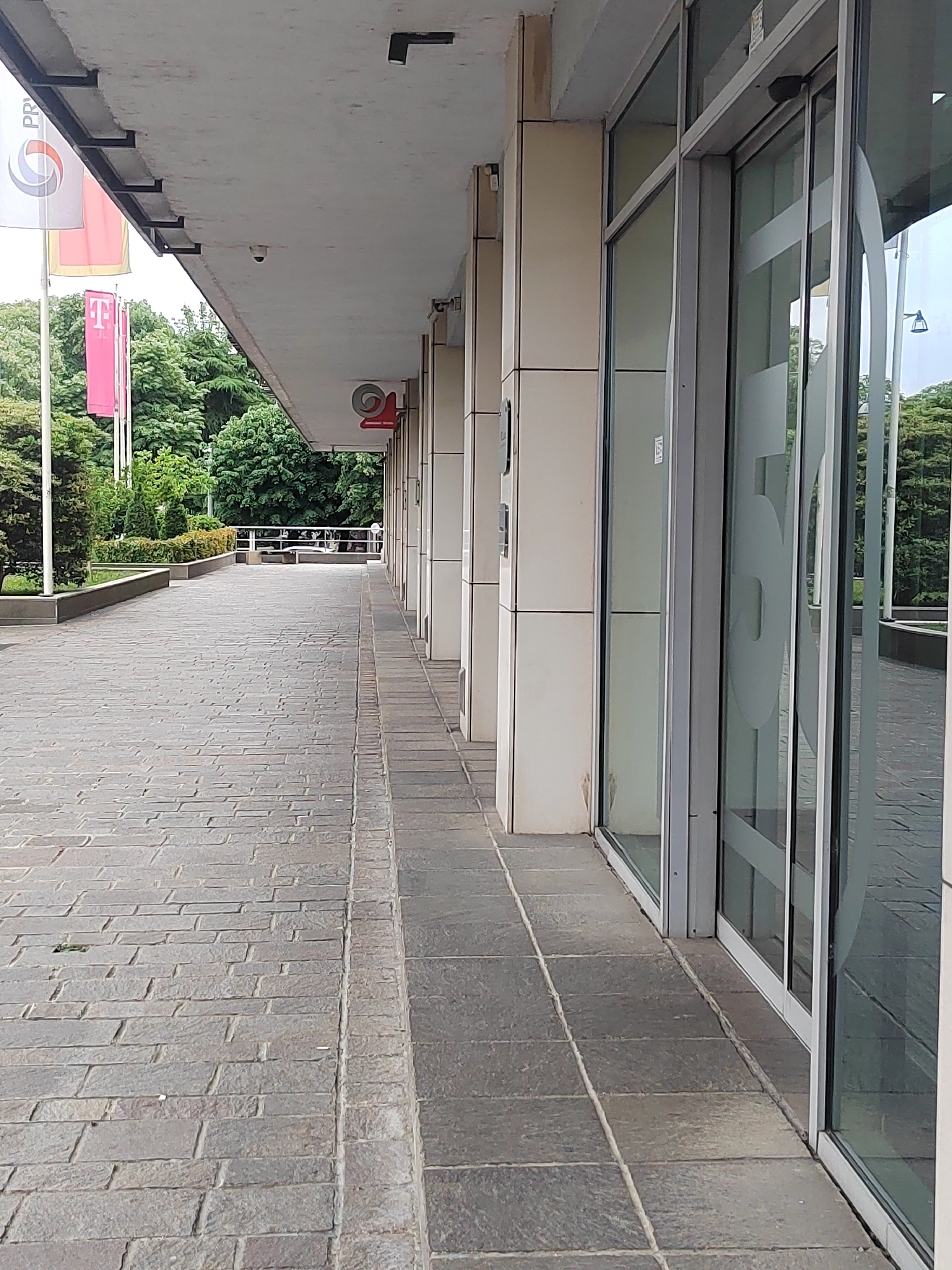 The image shows a covered walkway leading to the entrance of a building in Podgorica, Montenegro. The walkway is paved with gray stone blocks and is lined with rectangular support pillars. On the left side of the walkway, there are flags displaying company logos. Beyond the walkway, there are trees and greenery. The building's entrance features glass sliding doors with the numeral "50" etched on the glass.