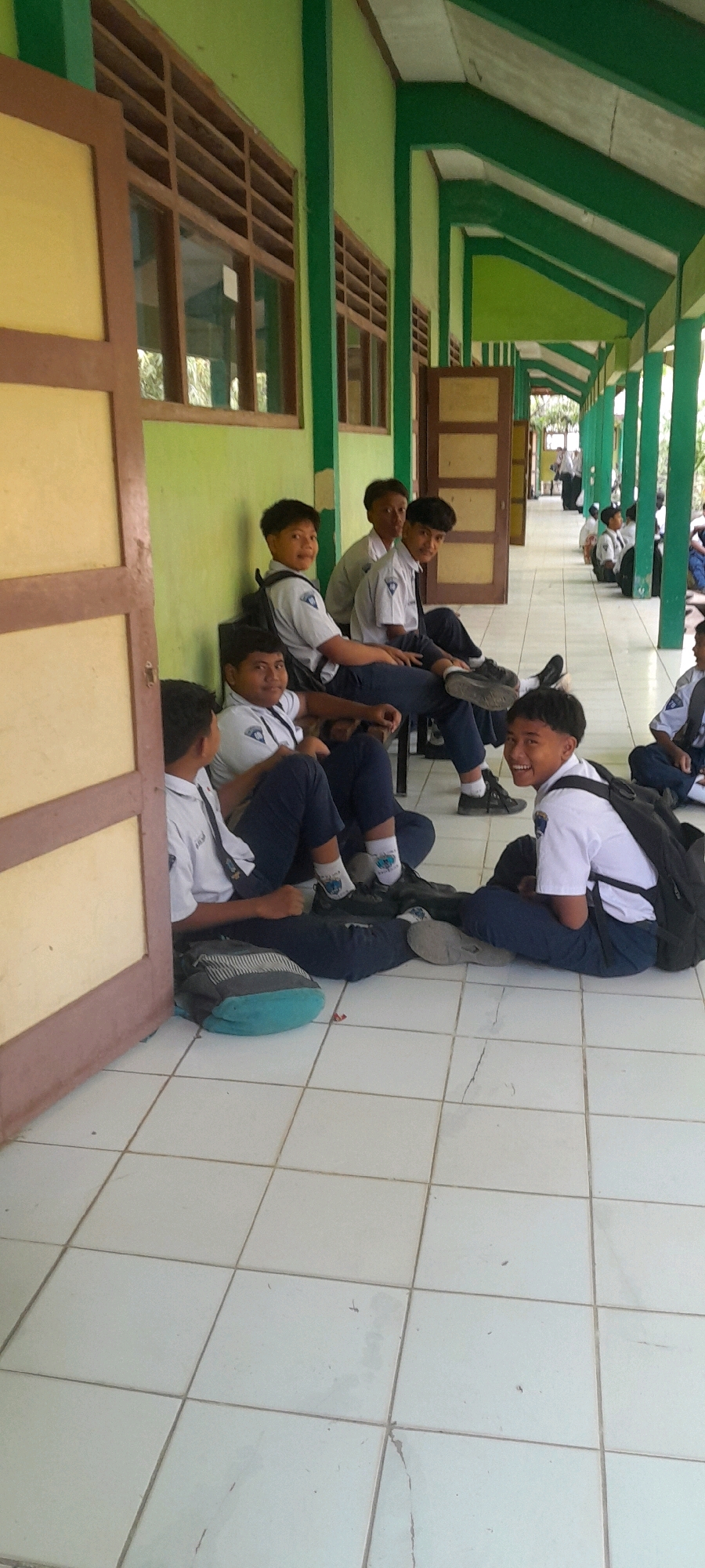 The image depicts a group of male students in school uniforms sitting on the floor and on chairs in a covered outdoor walkway or hallway. The walkway has a green painted ceiling and green columns. There are windows with wooden louvers on the left side of the walkway. The floor is tiled with light-colored square tiles.
In the foreground, on the left, a student in a white shirt and dark pants sits on the floor with a backpack next to him. To his right, another student sits on the floor, also in a uniform, looking towards the camera. Further back, a group of students are seated on chairs in the walkway. The student on the far right of this seated group is looking directly at the camera and smiling. He is wearing a white shirt, dark pants, and a backpack. In the background, other students are visible further down the walkway, some sitting and others standing. The overall impression is of students gathered in an informal setting, possibly during a break or waiting for something. The location is identified as Tahunan, Indonesia.