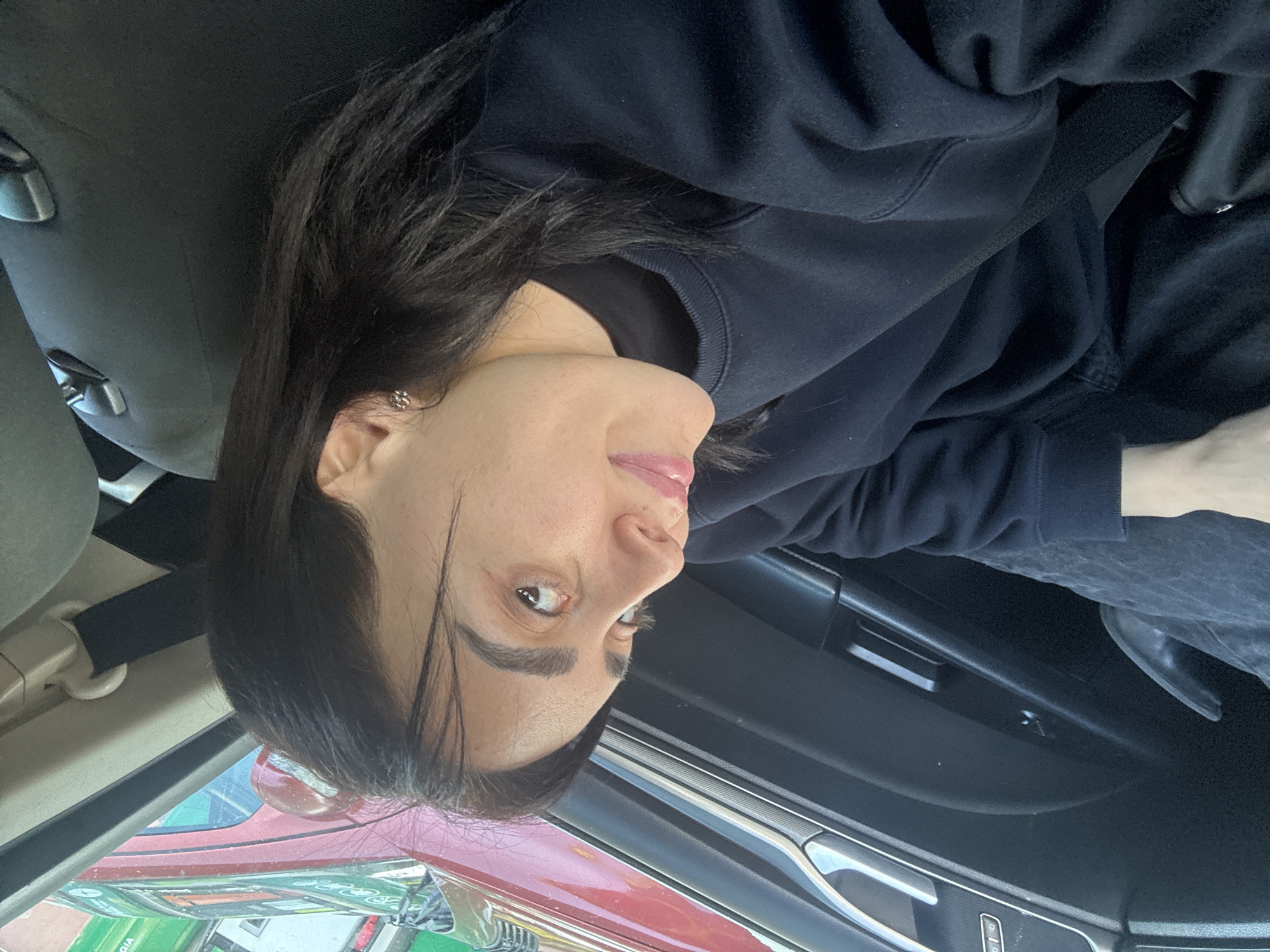 A woman with dark hair is wearing a dark blue sweatshirt and a seatbelt in the driver's seat of a car. The car is parked on a street in Tbilisi, Georgia, where red and green vehicles are visible outside.