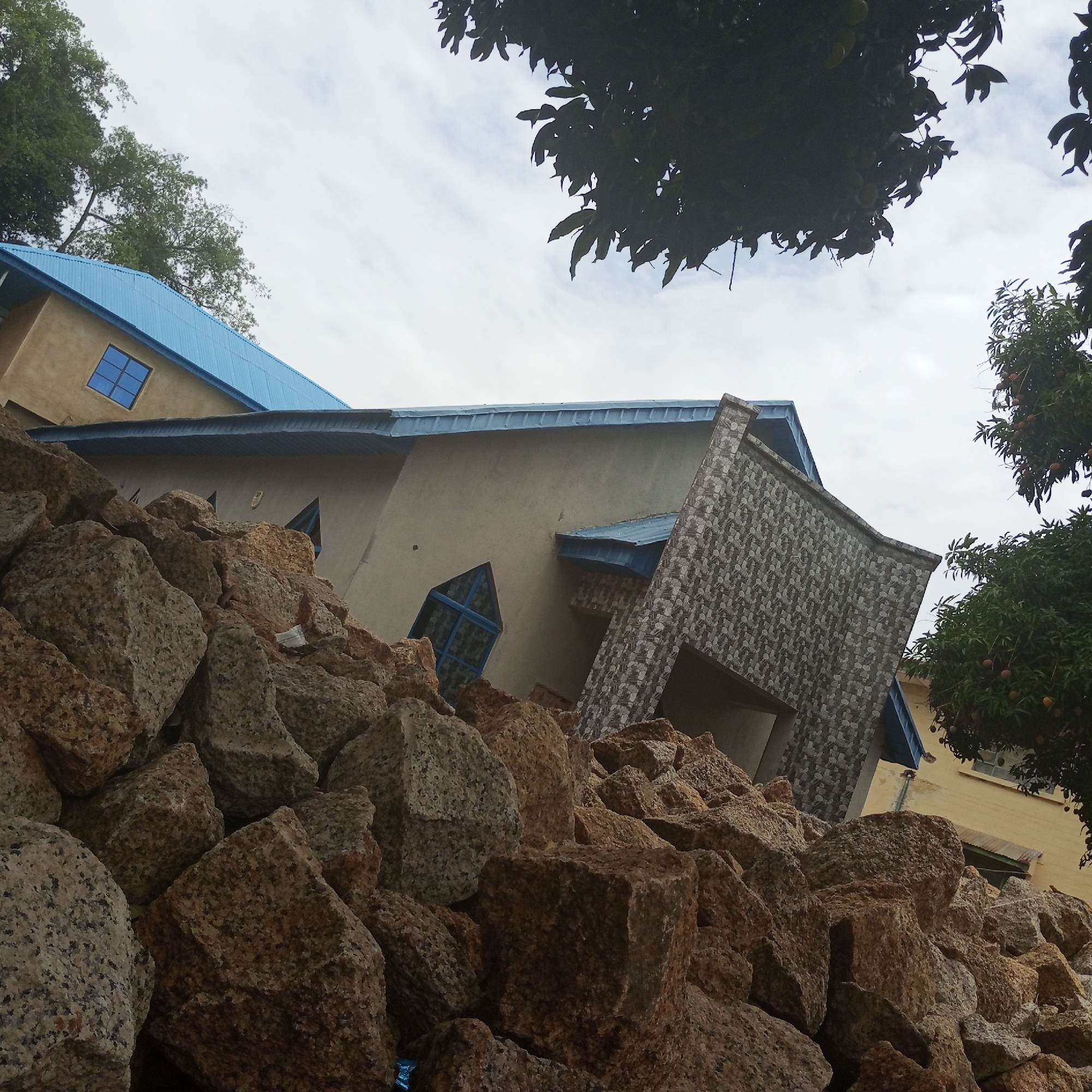 The image shows a pile of light brown and grey rocks in the foreground. Behind the rocks is a building with a beige exterior. The building features a blue corrugated metal roof, blue-framed windows, and a section of the facade covered in grey-patterned tiles. Two trees are visible, one with dark green foliage at the top of the image and another with lighter green foliage and reddish-brown fruit on the right side. The sky is overcast with light grey clouds. The building is located in Jos, Nigeria.