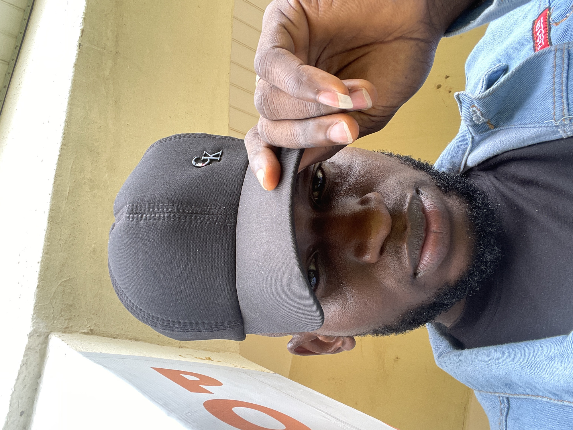 A close-up, low-angle shot shows a dark-skinned man wearing a black baseball cap and a light blue denim jacket over a black t-shirt. He has a short beard. The man is holding the brim of his cap with his right hand, partially obscuring his right eye. A silver logo, resembling the letters "CK" or "G K", is visible on the front of the cap. The background consists of a textured, yellowish wall and a portion of a white wall with orange lettering on the left side. The setting appears to be outdoors, possibly in the city of Durumi, Nigeria.