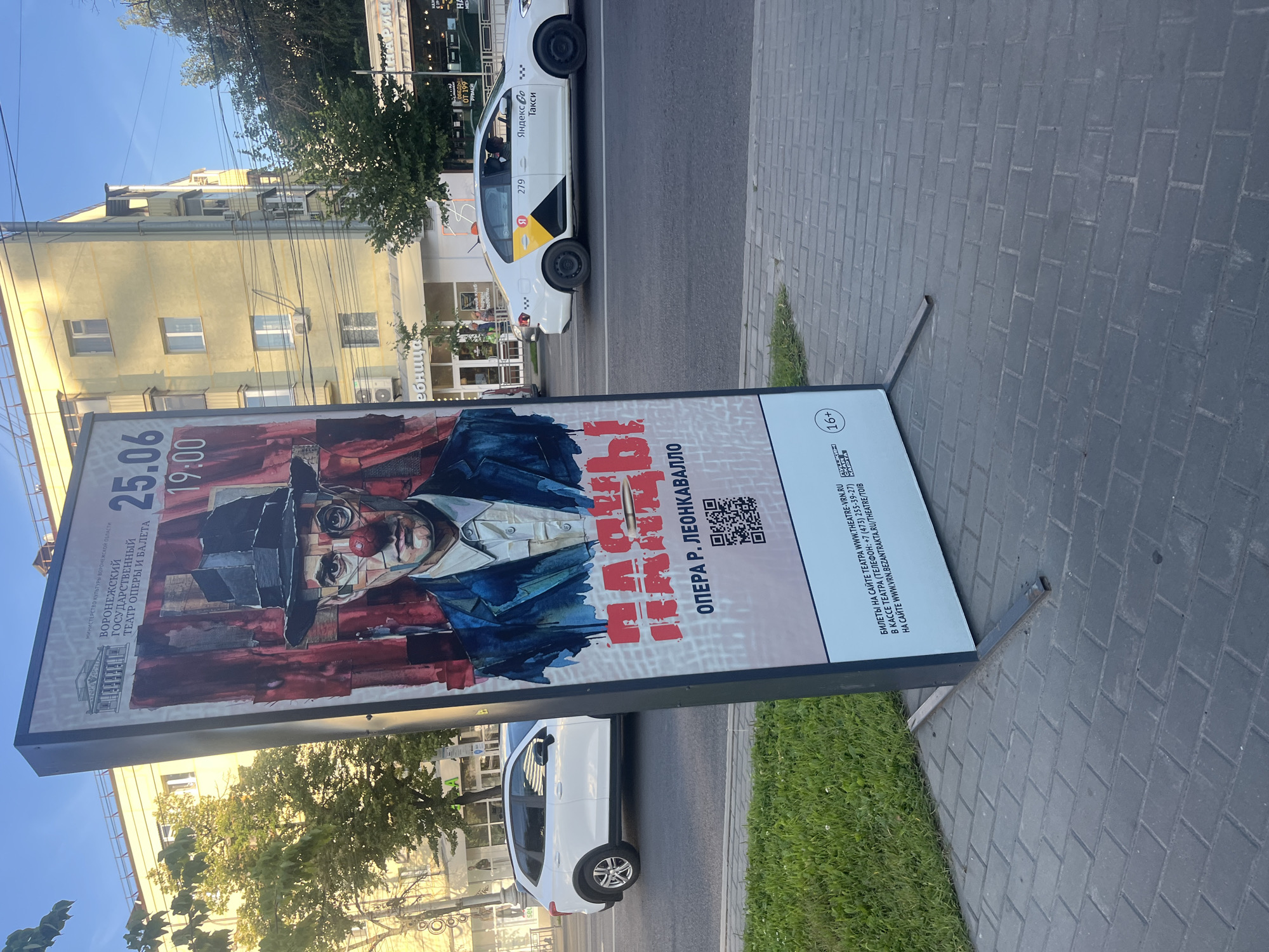This media file captures a street scene in Novaya Usman, Russia. The primary subject is a large outdoor advertising stand displaying a poster for an opera performance. The poster features a striking image of a person in a hat and red clown nose, with bold red text that appears to announce the opera "Pagliacci" (Паяцы). Visible text on the poster indicates the performance date as June 25th and the time as 19:00, along with the "Voronezh State Opera and Ballet Theatre" and other details like websites and phone numbers. An age restriction symbol "16+" is also present.
In the background, a white Yandex Taxi is visible, with its branding clearly displayed. The scene is set on a paved sidewalk adjacent to a street. Beyond the sidewalk and street, buildings and trees line the area, suggesting an urban or semi-urban environment. The lighting indicates it is daytime, likely late afternoon or early evening, given the long shadows and warm tones. The overall mood is neutral, focusing on the advertising and the urban setting.