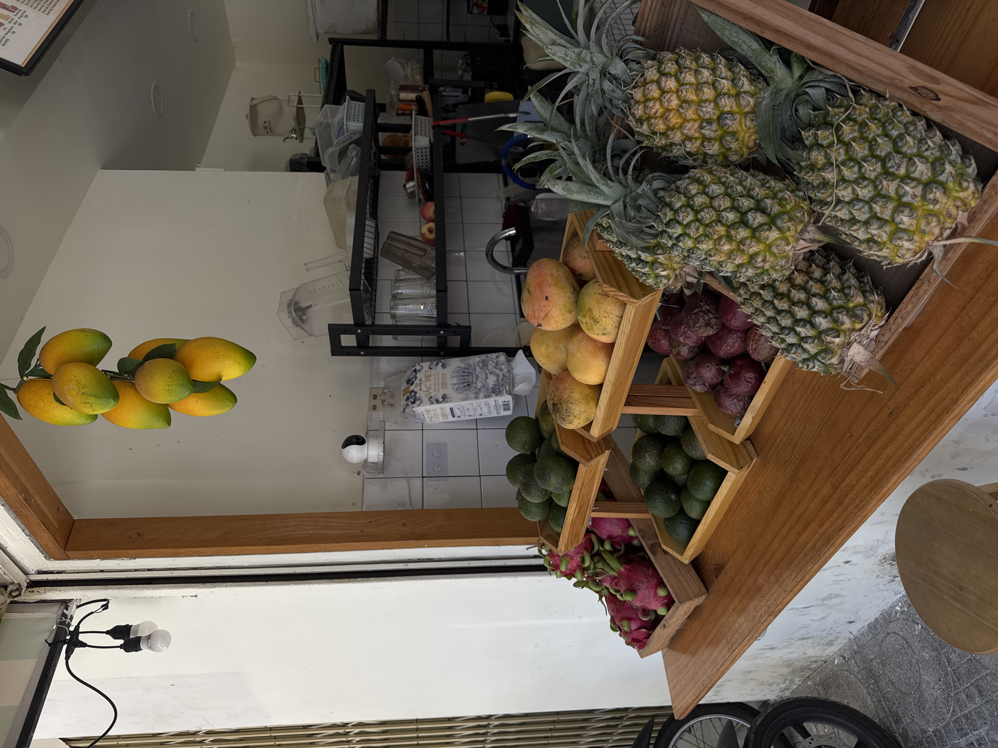 This is a photograph of a fruit stand, likely in Nha Trang, Vietnam, given the context. The scene is brightly lit, suggesting it's daytime. The fruit stand is made of wood and displays a variety of fresh fruits. Prominently featured are pineapples at the top, followed by mangoes and what appear to be passion fruits. Below these, avocados and dragon fruits are arranged in wooden crates. The background reveals the interior of a shop or stall, with shelves stocked with various items, including what might be beverages and kitchenware. A sink and a tiled wall are also visible, indicating a functional preparation area. A security camera is mounted on the wall. In the foreground, a portion of a street or walkway is visible, with the wheel of a motorcycle or bicycle. The overall impression is one of a vibrant, fresh produce offering.