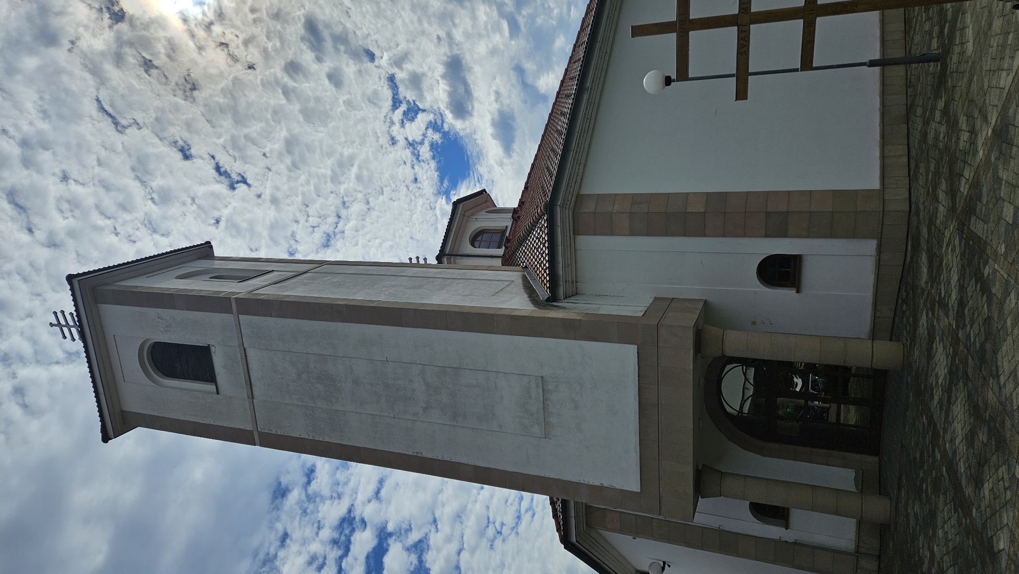 The image shows the exterior of a church, likely St. Nicholas Cathedral, located in Prešov, Slovakia. The architectural style appears to be a mix of modern and traditional elements, with a prominent bell tower. The building is painted white with beige accents, and a red-tiled roof is visible. In the foreground, a wooden cross stands next to a lamppost. The sky is partly cloudy, with a sunbeam breaking through the clouds on the right side of the frame. The ground in front of the church is paved with interlocking stones. No people are visible in the image.