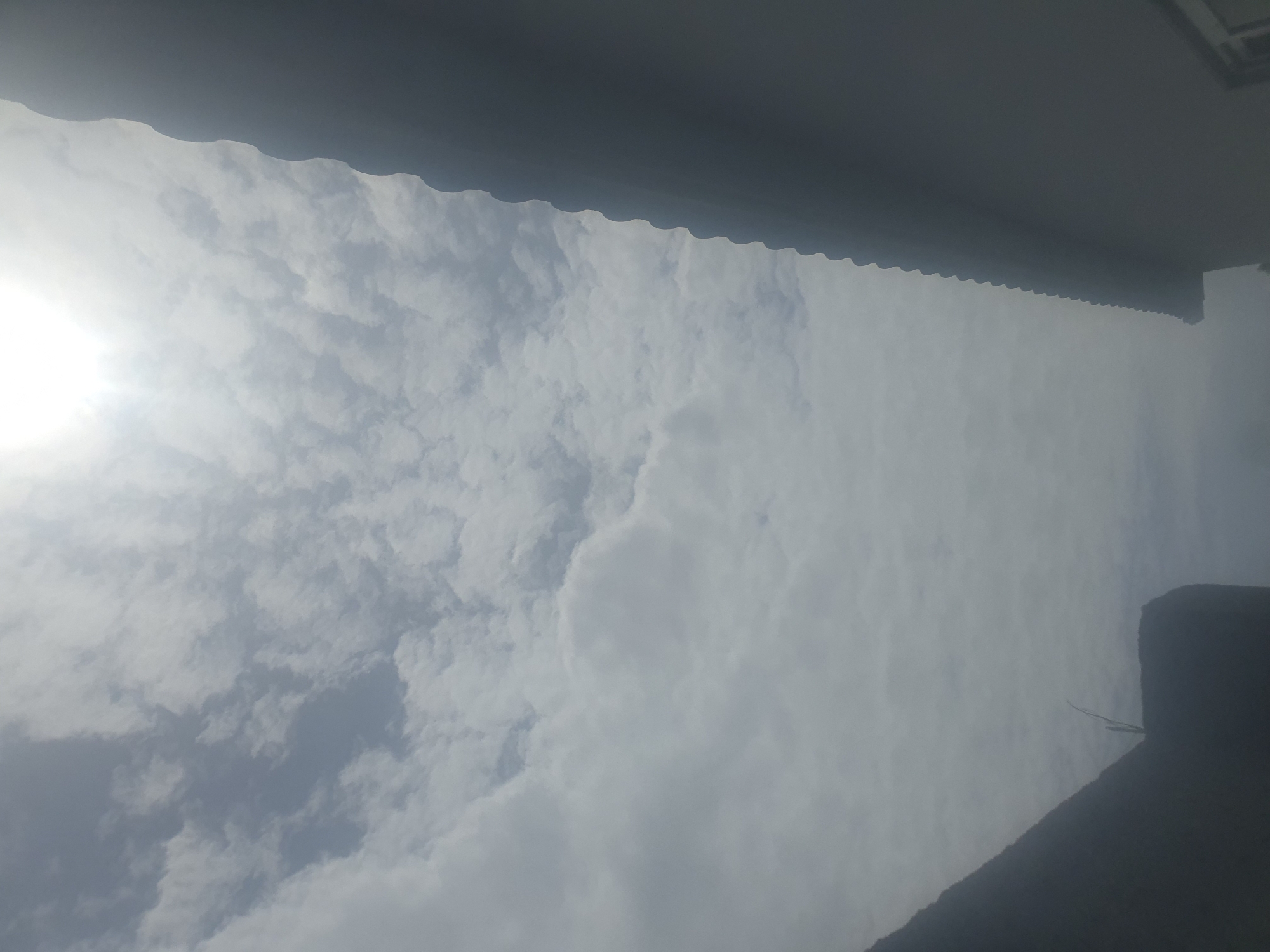 The image shows a sky filled with clouds, with the sun shining brightly on the left side. The sky appears to be overcast with scattered clouds, suggesting it might be a partly cloudy day. In the foreground, a dark, shadowed structure, possibly a roof or awning, is visible along the top and right edges of the frame, with a scalloped edge facing the sky. The perspective is from below, looking upwards. The time of day is likely daytime due to the bright sun. The weather appears to be bright but with cloud cover. The location context is Jos, Nigeria, but no specific landmarks are identifiable. There is no visible text in the image.