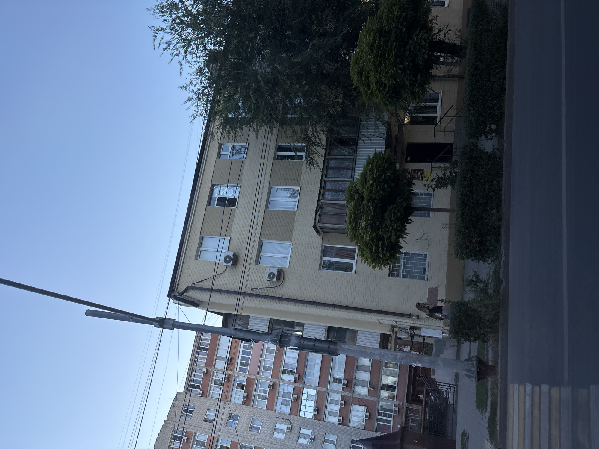 The image depicts a residential area in Rostov-on-Don, Russia. In the foreground, a street with a pedestrian crossing is visible. Behind the street, a light-colored, multi-story apartment building with several windows and air conditioning units is prominent. To the left of this building, a taller, red-bricked apartment building with numerous balconies and windows is partially visible. Trees and bushes line the sidewalk in front of the buildings. A utility pole with overhead wires stands between the two apartment buildings. A woman in a dark dress and carrying a bag is walking on the sidewalk in front of the light-colored building. The sky is clear and blue.