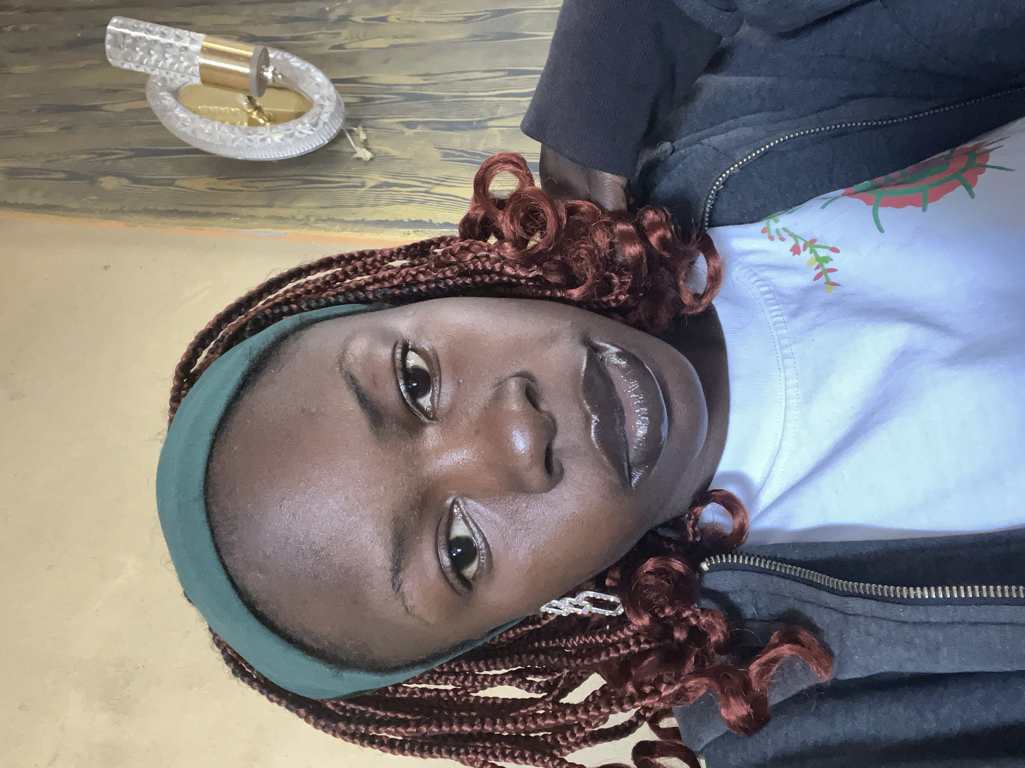 A close-up, head-and-shoulders selfie of a young Black woman with dark skin and braids that are reddish-brown in color. She is wearing a dark gray zippered hoodie over a white t-shirt with a floral pattern. She has on a dark green headband. Her lips are glossy and dark. She is wearing a silver earring in her left ear. The background is a wall with a patterned texture that appears to be wood grain and a brass and crystal light fixture mounted on the wall to her right. The lighting suggests an indoor setting, possibly a home, in Jos, Nigeria.