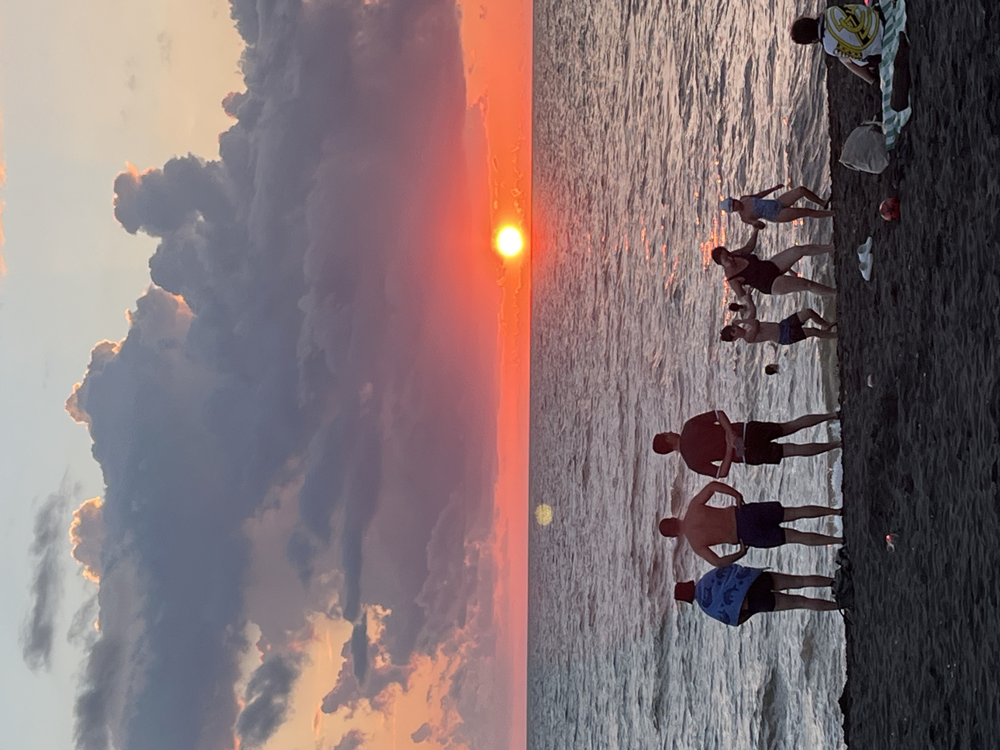 In Ureki, Georgia, a picturesque sunset bathes a sandy beach in warm, orange hues. The sky is a dramatic canvas of swirling clouds, transitioning from soft grays and purples to vibrant pinks and oranges near the horizon. The sun, a brilliant orb, sits low, casting a shimmering reflection on the calm sea.
On the dark, sandy shore, a group of people are gathered. In the foreground, a lone figure in a black and yellow patterned shirt sits on a striped towel, facing away from the camera. To their left, a red ball and a light-colored bag rest on the sand.
Further down the beach, closer to the water's edge, a family enjoys the evening. A woman in a dark swimsuit holds the hands of a young boy and a girl in a light blue swimsuit as they appear to be playing or dancing. Behind them, several other individuals stand and observe the scene. Three men are positioned together, two facing the sea and one turned slightly towards the others. One man is wrapped in a blue towel, while another stands with his hands behind his back. A third man, wearing a dark shirt and shorts, also faces the water. The sea itself is a gentle expanse of gray-blue, with small waves lapping at the shore.