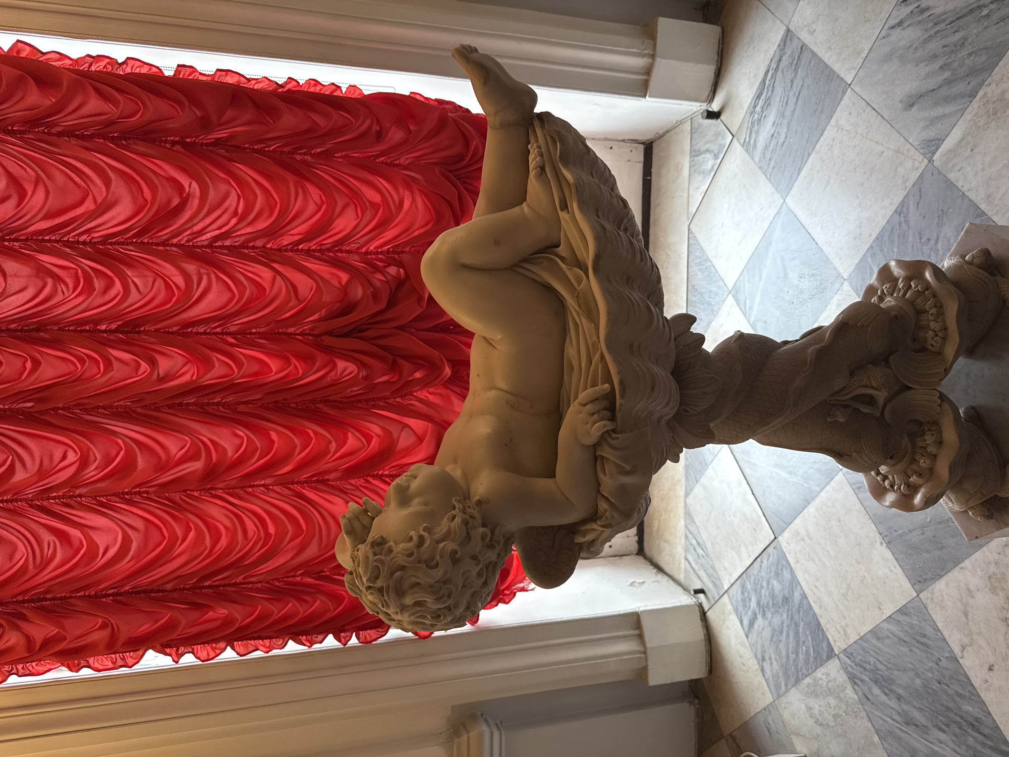 The image shows a marble sculpture of a cherub reclining on a shell, which is supported by a pedestal. The sculpture is placed on a floor tiled in a checkered pattern of light and dark marble. Behind the sculpture, a vibrant red curtain is draped, creating a dramatic backdrop. To the left of the sculpture, a white fluted column is visible, suggesting an interior setting. The overall scene evokes a sense of classical art and opulence. This sculpture is likely located in the Catherine Palace in Pushkin, Russia, a place renowned for its exquisite art collection and grand architecture.