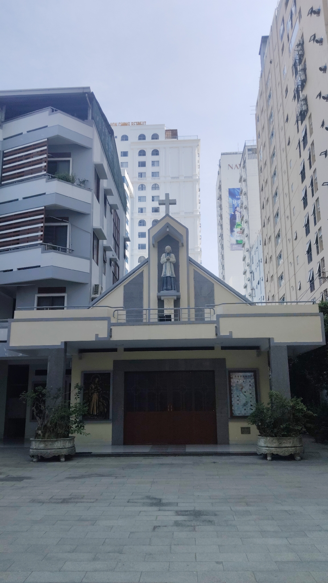 The image shows a church facade in Bac Nha Trang Ward, Vietnam. The church features a central statue of a saint beneath a cross, above a dark-colored entrance door. Artwork or signage is displayed on either side of the door. The surrounding area is paved with gray blocks. The church is flanked by modern buildings; one is a grey building with horizontal brown slats. Taller buildings rise behind, including one with “ION CUONG RETREAT” on top. Another one has a tall advertisement on it. Potted plants sit on both sides of the church entrance. The sky is visible at the top. There is nobody visible in the image.