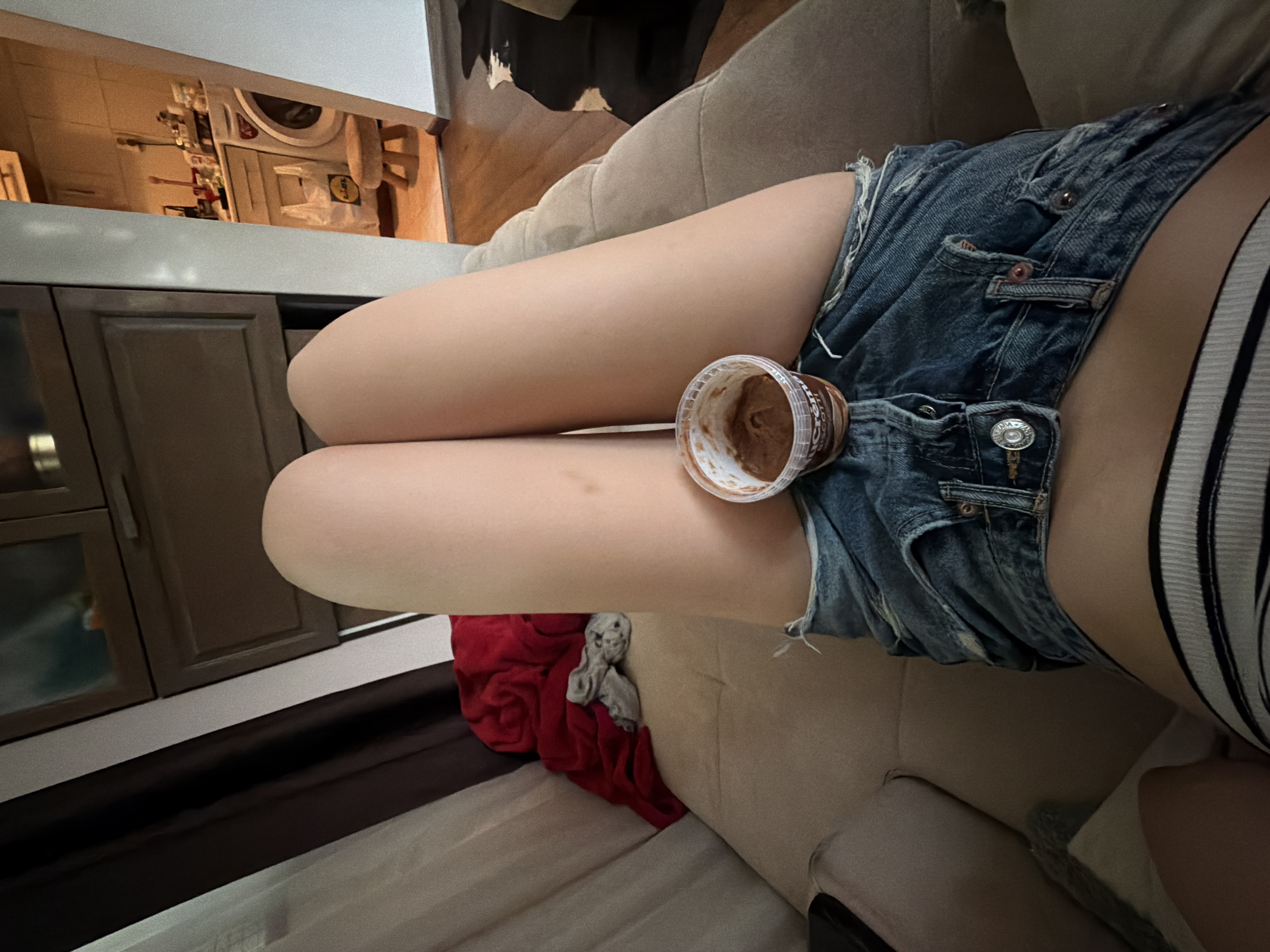 The image presents a first-person view looking down at a person's bare legs, clad in distressed denim shorts and striped underwear, resting on a light beige sofa. A small, clear plastic container of a brown spread, partially labeled "NuCrem Biscotti," is positioned between the person's thighs. Minor marks are visible on the right thigh. To the immediate left on the sofa, a pile of red and grey fabric is present. The background features dark brown curtains and a wooden cabinet with glass-fronted doors. Through an opening on the right, an adjacent room, likely a kitchen or utility area, is visible. This room contains a white washing machine, a small wooden three-legged stool, and a white "Lidl" branded shopping bag on a wooden floor. Various kitchen items are visible on a counter in the background. A dark, possibly patterned rug, is partially visible on the floor in the background room. The scene is an indoor residential setting, presumed to be in Tsarevo, Bulgaria.