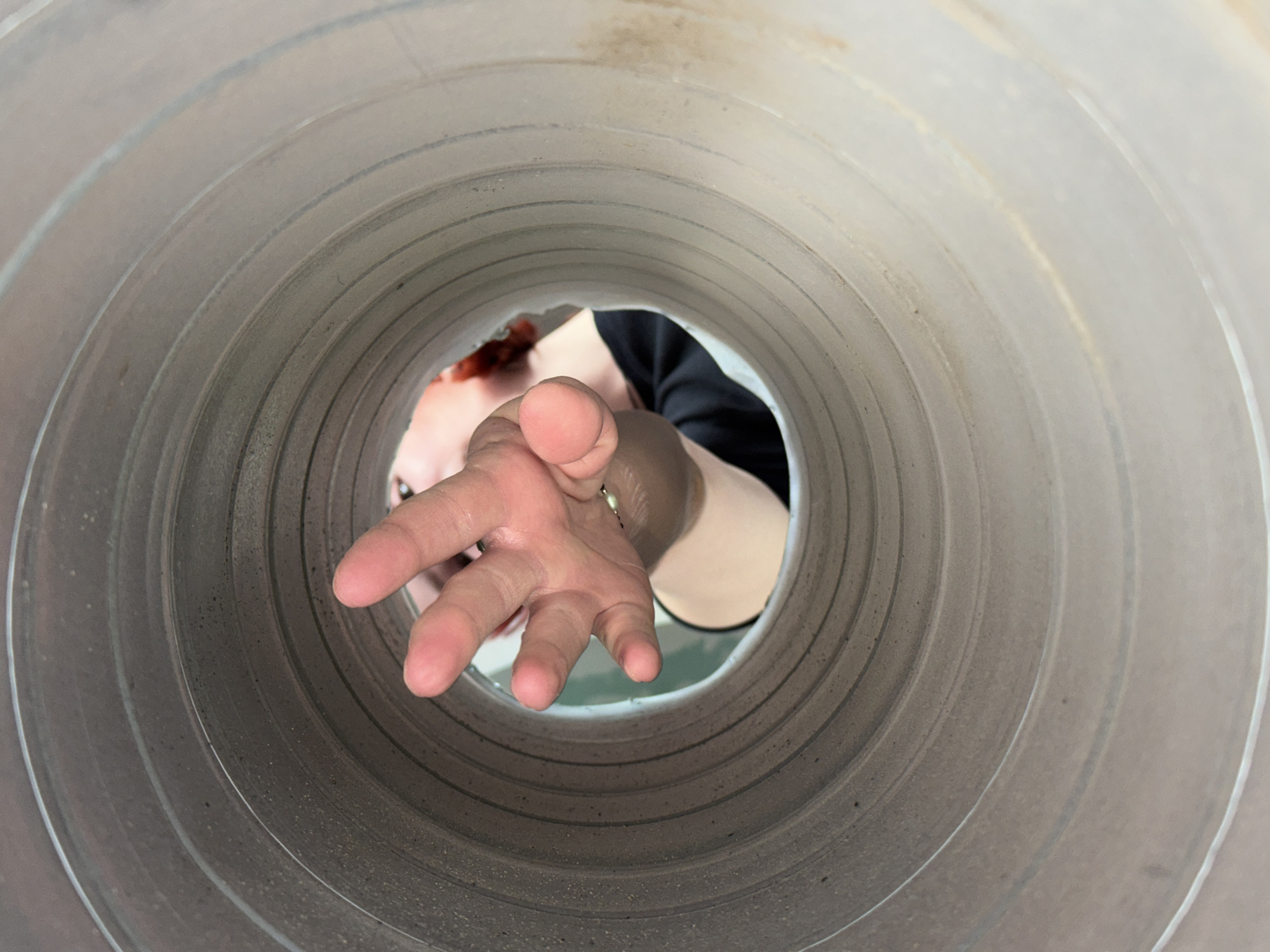 A low-angle shot looking up through the interior of a large, corrugated, cylindrical pipe. A person is looking into the pipe with their hand extended, fingers outstretched. The person appears to have red hair and is wearing a black top. A silver bracelet is visible on their wrist. The pipe is gray and has multiple concentric ridges. The location is unknown.