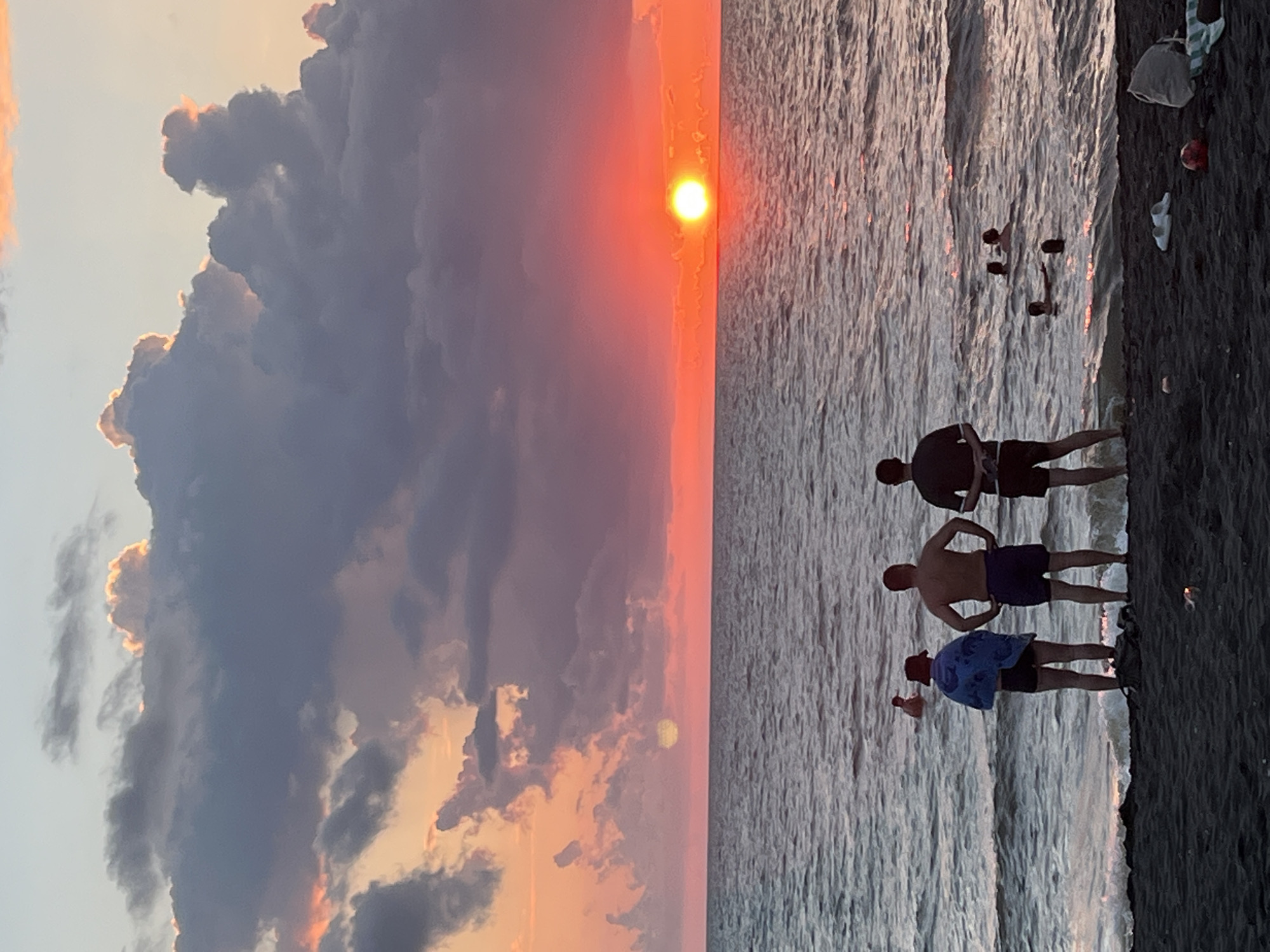 The image depicts a sunset over the Black Sea in Ureki, Georgia. Three individuals stand on a dark sand beach, facing the water. The person on the left wears shorts and a blue towel draped over their shoulders, and a hat. The middle person wears shorts and has their hands on their hips. The third person, on the right, wears shorts and has their arms crossed. Several other people are visible in the water, swimming. The sky is filled with dramatic clouds, tinted with hues of orange, pink, and purple, as the sun sets on the horizon, casting a vibrant glow on the water. The sea is choppy with moderate waves. Various items are scattered on the beach, including a bag, a pair of flip-flops, and a ball.