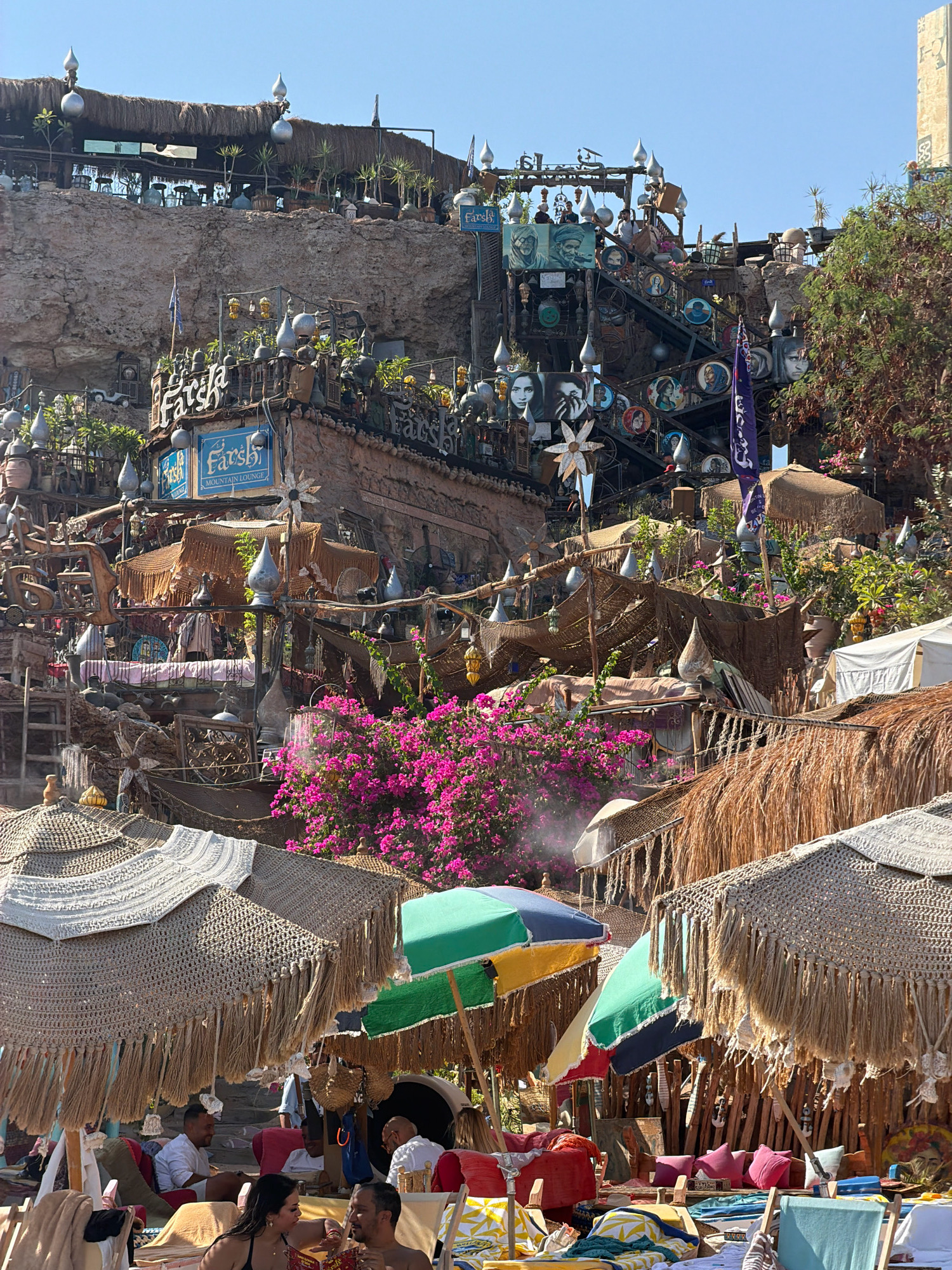 This image captures a vibrant outdoor scene, likely a beach resort or lounge area, at Farshta Mountain Lounge in Sharm El Sheikh, Egypt. The architecture is built into a rocky mountainside, featuring multiple levels adorned with numerous decorative elements, including lamps, hanging ornaments, framed portraits, and flags. The structures are covered with straw and natural materials, creating a rustic and bohemian aesthetic.
In the foreground, several beach umbrellas with woven tops and fringed edges provide shade over lounge chairs and seating areas. Brightly colored beach umbrellas, some with multi-colored panels, are also visible. People are seated on lounge chairs, with some engaged in conversation. A couple in the lower foreground is looking at each other, with one person gesturing towards the other. Other individuals are seated or lounging in the background, enjoying the atmosphere.
The environment is characterized by abundant greenery, including lush pink bougainvillea flowers cascading down the structures. The sky is clear blue, indicating a sunny day. Various objects, such as cushions, towels, and decorative items, are scattered around the seating areas, contributing to the relaxed and inviting ambiance. The overall impression is one of a lively and picturesque recreational space.