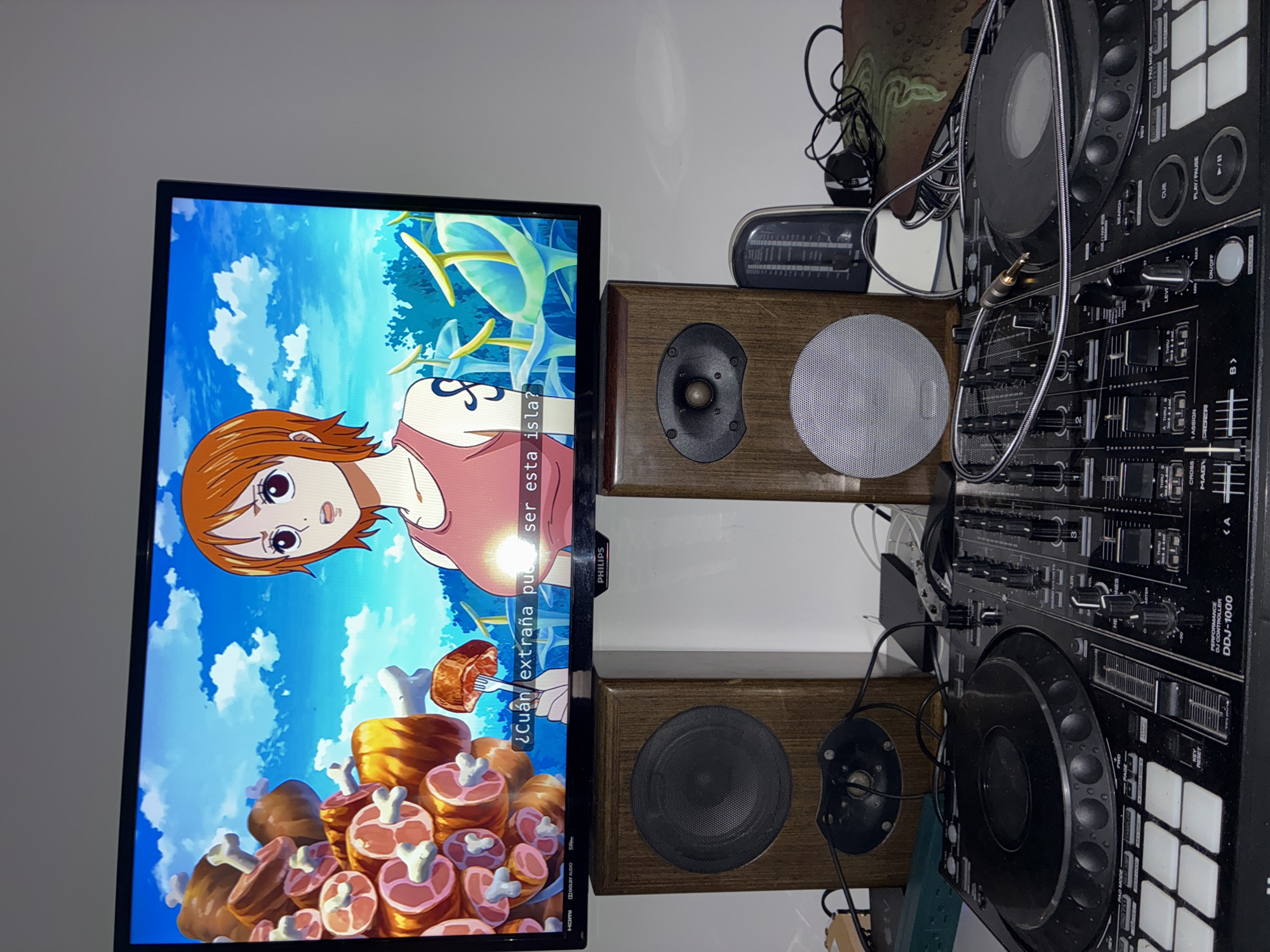 This image shows a television screen displaying a scene from the anime "One Piece," featuring Nami standing in front of a sky background and a large pile of meat. The television is located next to two wooden speakers and a black Pioneer DJ DDJ-1000 DJ controller. The scene appears to be indoors, and the specific location is Huixquilucan de Degollado, Mexico.