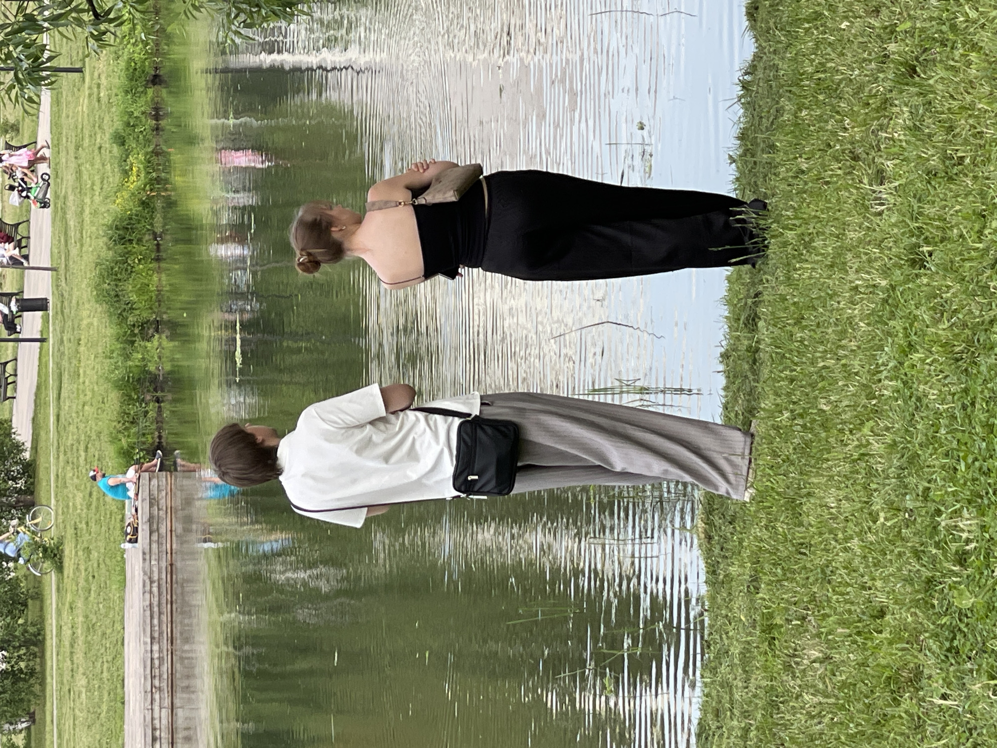 Two individuals, a male and a female, stand on the grassy bank of a body of water. The male, facing away from the camera, wears a white collared shirt and gray striped pants, with a black crossbody bag slung over his right shoulder. The female, also facing away, is dressed in a black tank top with thin straps and loose-fitting black pants, carrying a small beige purse over her left shoulder. Both appear to be looking out at the water. In the background, across the water, a wooden structure and more greenery are visible, along with distant figures and a person on a bicycle. The scene suggests a park setting, likely within Moscow, Russia, given the presence of typical urban park elements and landscaping.