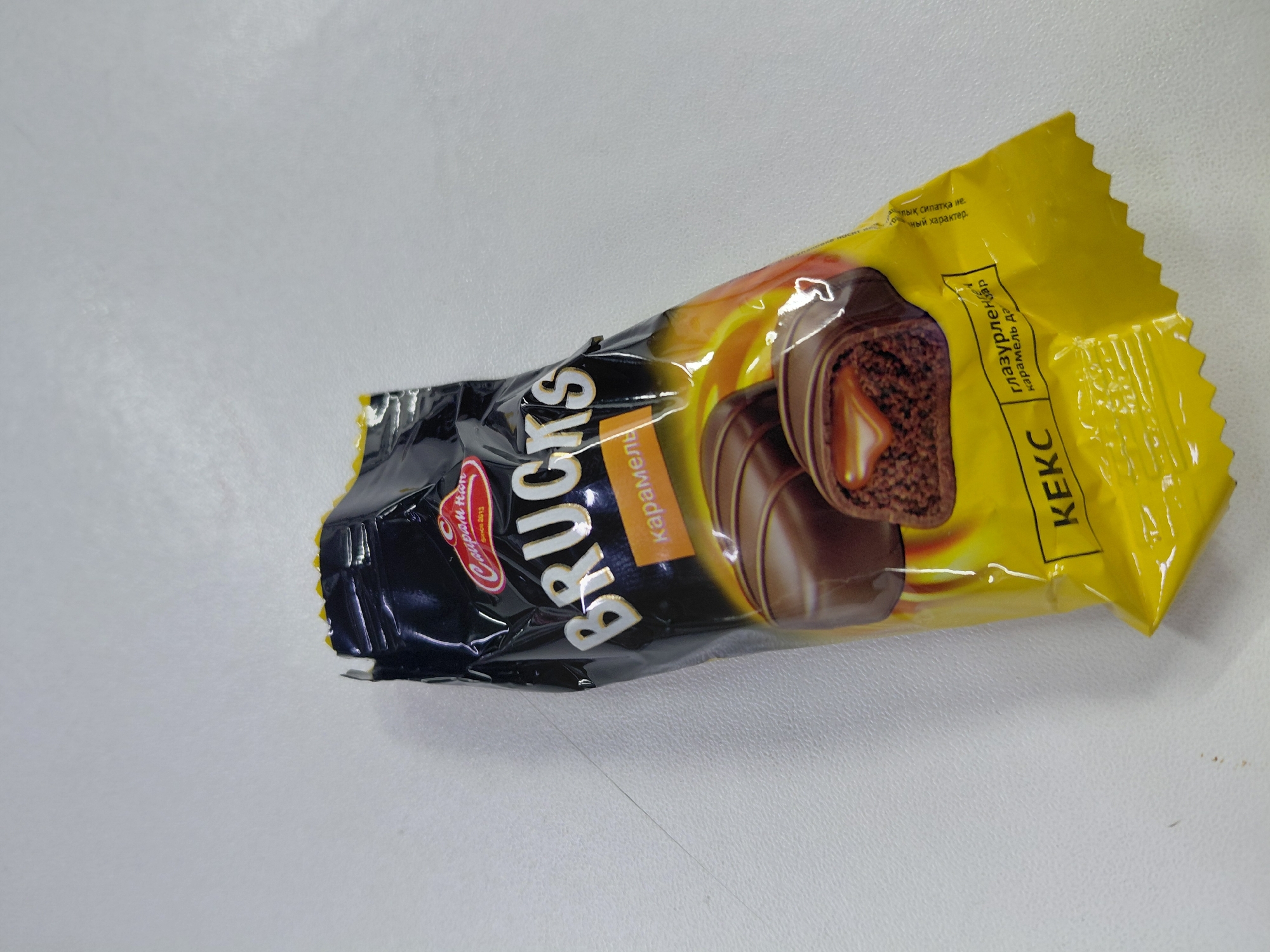 The image shows a single, individually wrapped chocolate cookie with caramel filling. The wrapper is primarily black with yellow accents and features the brand name "BRUCKS" in large, gold lettering. The cookie itself appears to be chocolate-covered with a visible caramel center. The wrapper also displays text in what appears to be Cyrillic script. The item is placed on a white, textured surface, possibly a table or desk. There is no indication of any specific location in Aktau, Kazakhstan, or any people present in the image.