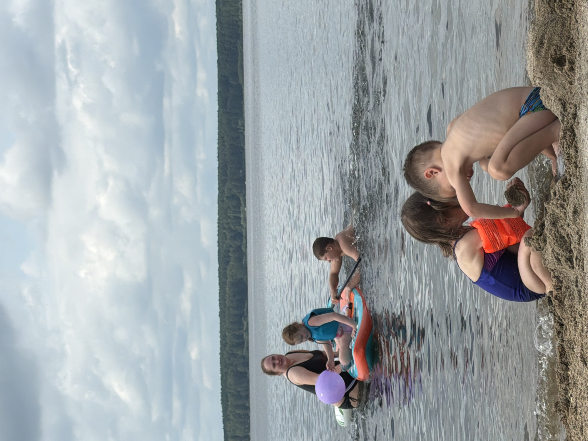 The image depicts a summer day at a lake, likely in Zarechny, Russia, where a family is enjoying recreational activities. In the foreground, two young children, a girl in a blue swimsuit with orange arm floaties and a boy in patterned swim trunks, are crouched on a sandy beach, engaged in playing with sand. The girl is holding a clump of wet sand. In the background, a woman in a black swimsuit and a young girl in a blue life vest are in the water, sitting on an inflatable raft. A teenage boy is in the water beside the raft, holding a paddle and swimming. The lake is calm, with a tree line visible on the horizon. The sky is overcast with white clouds.