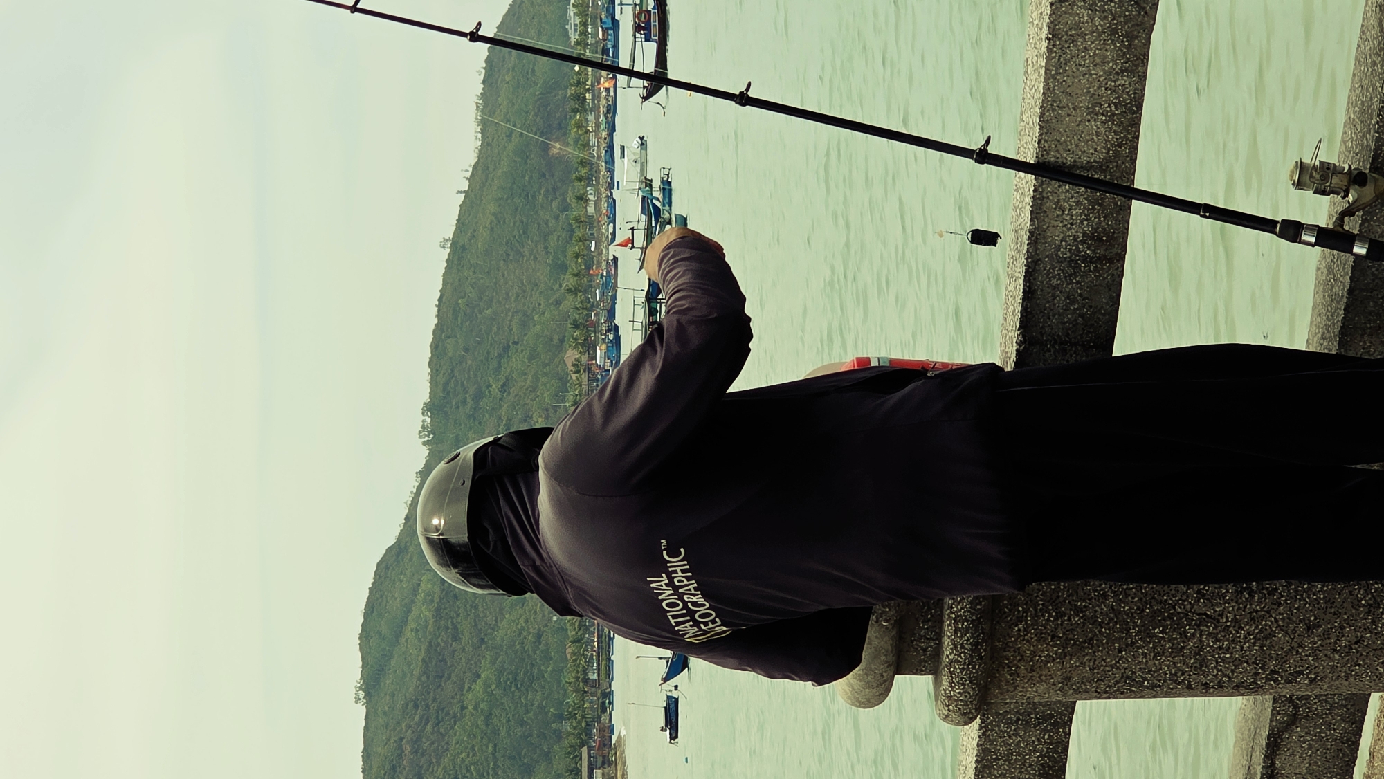A person wearing a black motorcycle helmet and dark clothing is fishing from a concrete structure. The person is holding a fishing rod over water, with a fishing bobber visible in the water. A hill covered in green trees is visible in the background, along with boats in the water. The location is Bac Nha Trang Ward, Vietnam.