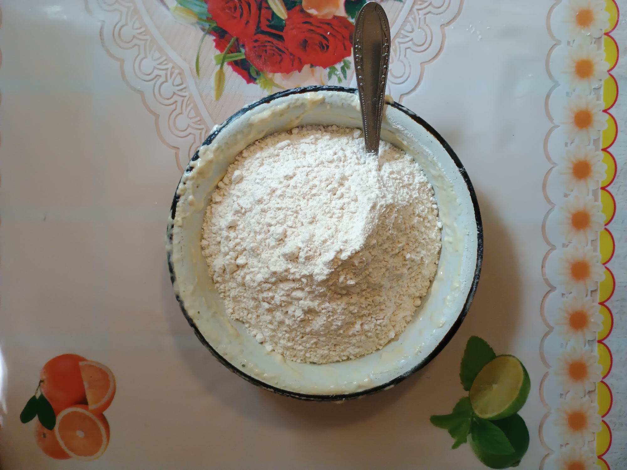 The image depicts a close-up, overhead view of a bowl filled with flour, resting on a table with a decorative floral border. To the left of the bowl, a spoon is partially submerged in the flour. The table also features a depiction of sliced oranges and two limes with leaves, positioned near the bottom left corner. In the background, to the right of the bowl, a floral pattern with red roses is visible. The scene is lit from above, casting a slight shadow from the bowl onto the table. No people are present, and no specific location in Zaporizhzhia, Ukraine, can be identified from the visual information provided. The overall setting appears to be a domestic kitchen or dining area.