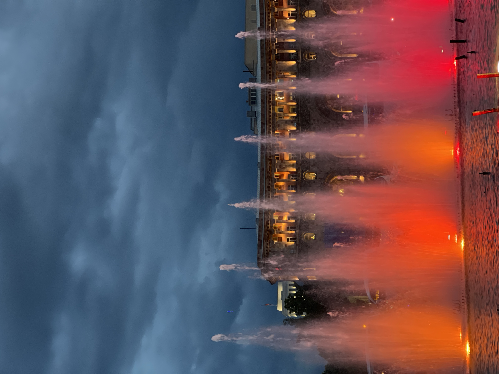 The image depicts the Republic Square in Yerevan, Armenia, at night. In the foreground, a fountain display is illuminated with red and orange lights. Streams of water shoot upwards, partially obscuring the view of the buildings in the background. The sky is overcast with dark, stormy-looking clouds. The buildings are lit up, casting a warm glow that contrasts with the dark sky. The image is rotated 90 degrees counterclockwise.