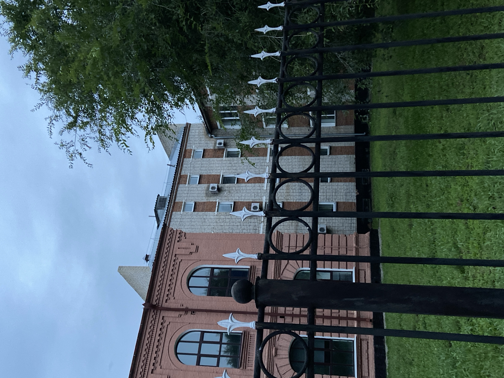 The image depicts a view of a building and its surroundings, seen through a black metal fence with decorative white finials and circular motifs. The building on the left is constructed with pinkish-red brick and features arched windows with dark frames. Adjacent to it, a section of the building is made of light-colored brick with red brick accents and features numerous rectangular windows and several air conditioning units mounted externally. A large green tree with dense foliage is visible on the right side of the image, partially obscuring the building. The ground in front of the fence is covered with green grass. The sky above is overcast with a dull, cloudy appearance. The scene suggests an urban or institutional setting, possibly an educational or administrative facility. The building's architectural style and the presence of air conditioning units are common in many regions, and without specific landmarks or identifying signs within the image, pinpointing an exact location within Blagoveshchensk, Russia, or any other city is not possible based solely on visual information.