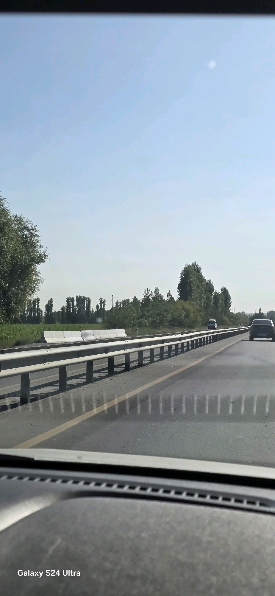The image is a photo taken from inside a vehicle looking out at a highway. The sky is clear and blue with a small white object visible in the upper right quadrant. A row of trees lines the right side of the highway, and a concrete barrier is visible to the left. The highway has a yellow line marking the lanes. Two cars are visible on the highway, one in the distance and one closer to the foreground. The bottom of the image shows the dashboard of the vehicle. A watermark in the bottom left corner reads "Galaxy S24 Ultra". The location is identified as Katput, Uzbekistan.