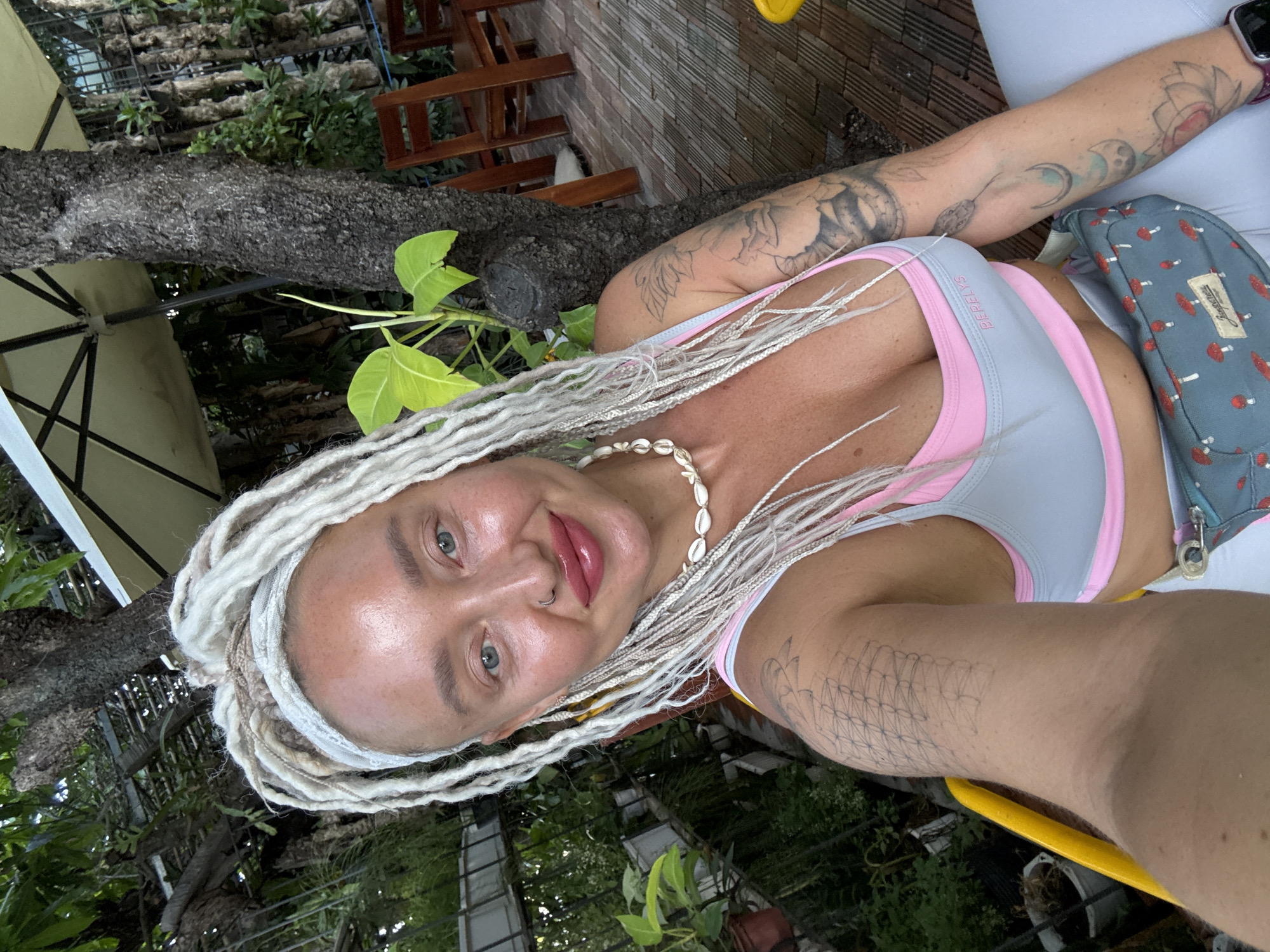 A woman with platinum blonde dreadlocks, a grey and pink sports bra, and a grey fanny pack with a mushroom print is taking a selfie outdoors. She is wearing a white shell necklace and a nose ring. Her arms are visible, displaying tattoos. The background suggests an outdoor seating area with trees, plants, wooden chairs, and a patio umbrella. The image is shot from a slightly upward angle. The location is specified as Nha Trang Ward, Vietnam.