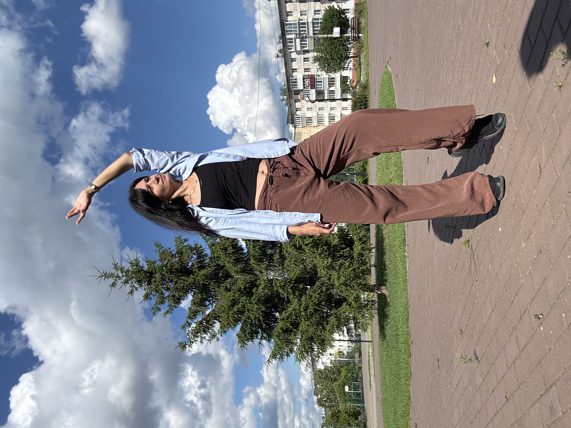 A full-body, outdoor, low-angle shot captures a woman with long, dark hair, dressed in a blue open button-down shirt over a black tank top and brown wide-leg pants, standing on a paved walkway in Chaikovsky, Russia. Her right arm is raised, palm facing upwards, as she gazes to her right. A tall evergreen tree stands behind her, with a large, fluffy cloud formation in the bright blue sky overhead. In the background, a multi-story residential building with white and beige facades is visible, along with a green lawn and park benches. The paved walkway is made of reddish-brown brick pavers, with small green plants emerging from cracks. The woman's shadow is cast on the ground to her right.