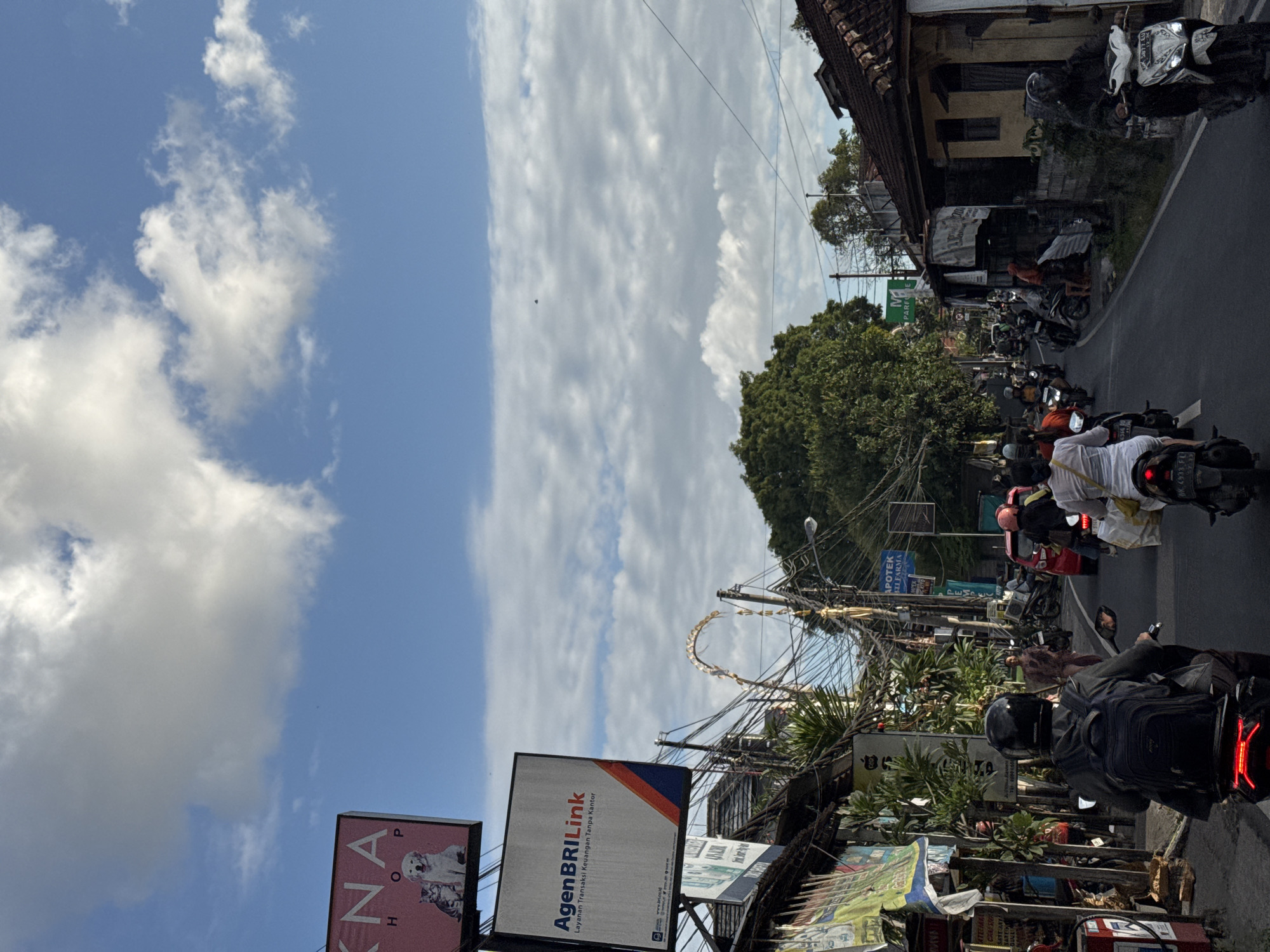 The image depicts a street scene in Denpasar, Indonesia, with a focus on motorbikes and pedestrian activity. The sky is partly cloudy, with a mix of blue sky and white clouds.
In the foreground, a person wearing a black helmet and a backpack is riding a motorbike. The motorbike's brake lights are illuminated. To the right, another person on a motorbike, also wearing a helmet, is visible from the side. The motorbike has a license plate that reads "DK 5877 EU". Further down the street, more motorbikes and some cars are present, indicating traffic.
Along the sides of the street, there are shops and buildings with various signs. One prominent sign is for "KNA SHOP" with images of a cat and a dog. Another sign reads "AgenBRIiLink". There are also numerous utility wires and poles, characteristic of urban infrastructure. A large tree with dense foliage is visible in the background, and a building with a dark roof and yellow walls is on the right side of the street.
The overall impression is a bustling street in a tropical urban environment, likely during daylight hours, with a focus on local commerce and transportation.