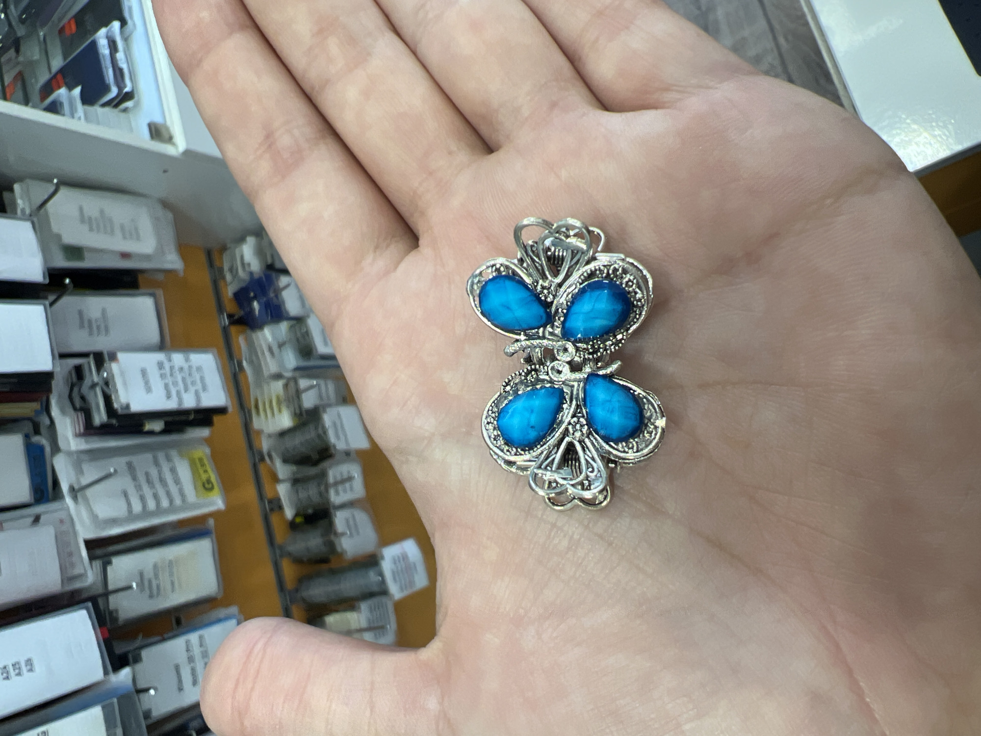 A close-up shot shows a person's open hand holding a decorative butterfly-shaped charm. The charm is silver-colored with intricate scrollwork and four bright blue gemstone-like inlays, arranged to resemble butterfly wings. The background is blurred, showing rows of small, individually packaged items on shelves, some with white labels and others with visible text. Among the visible text is a yellow label with the word "Gooo" in a stylized font and some smaller black text that is mostly illegible. The lighting suggests an indoor setting, possibly a store or market, given the display of merchandise. The setting appears to be Burgas, Bulgaria, based on the provided context. No specific time of day or weather conditions are discernible. The focus is on the hand and the charm, with the background providing context without being the primary subject. The hand is gently cupped, presenting the charm to the viewer.